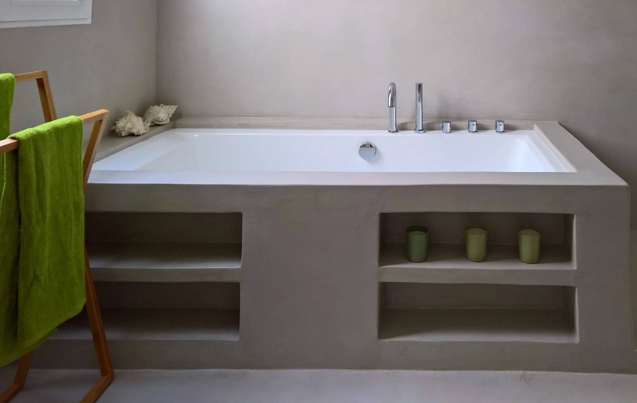 Bañera blanca empotrada en la pared con grifo y accesorios de metal, debajo hay estanterías con cuatro velas en vasos de vidrio y un juego de toallas verdes sobre una silla de madera a la izquierda.