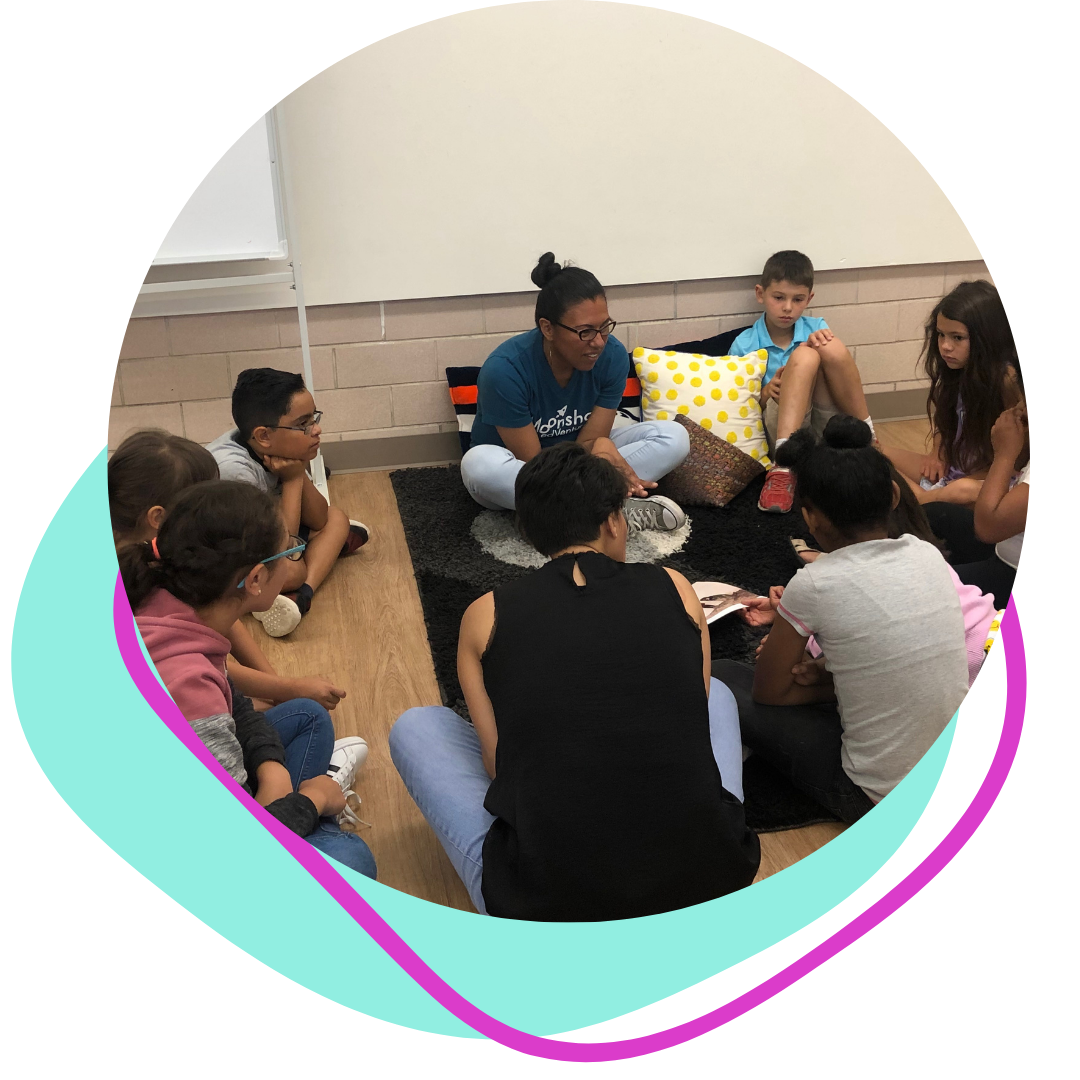 A woman sitting on a black rug with children gathered around her, engaged in a discussion or story time in a classroom setting.