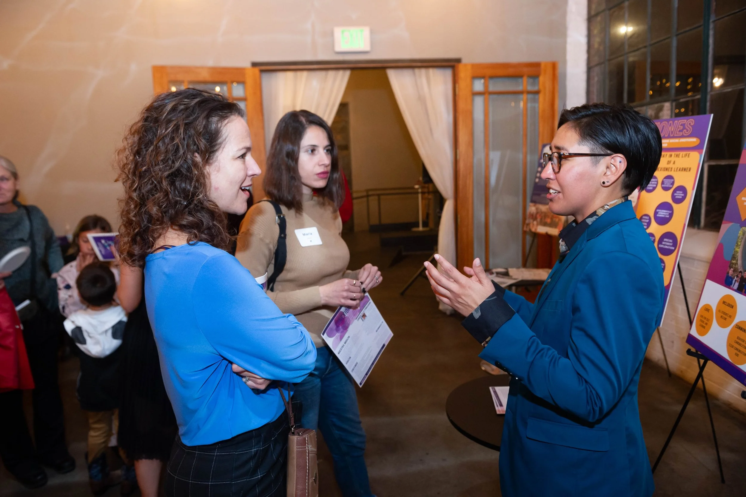 Three women engaged in conversation at an indoor event, with a poster display in the background.