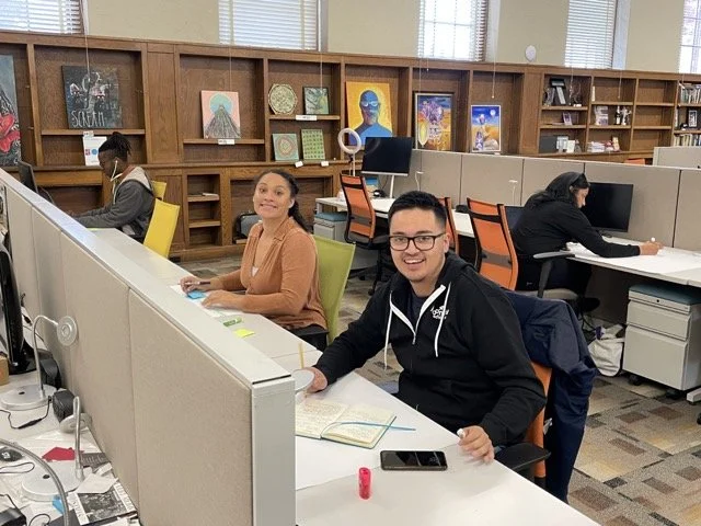 People working at computers in an office with wooden bookshelves displaying colorful artwork in the background.