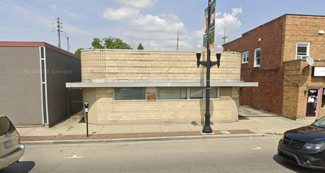 A small beige brick building with a flat roof, three narrow horizontal windows, located between a gray building and a red brick building on a city street.