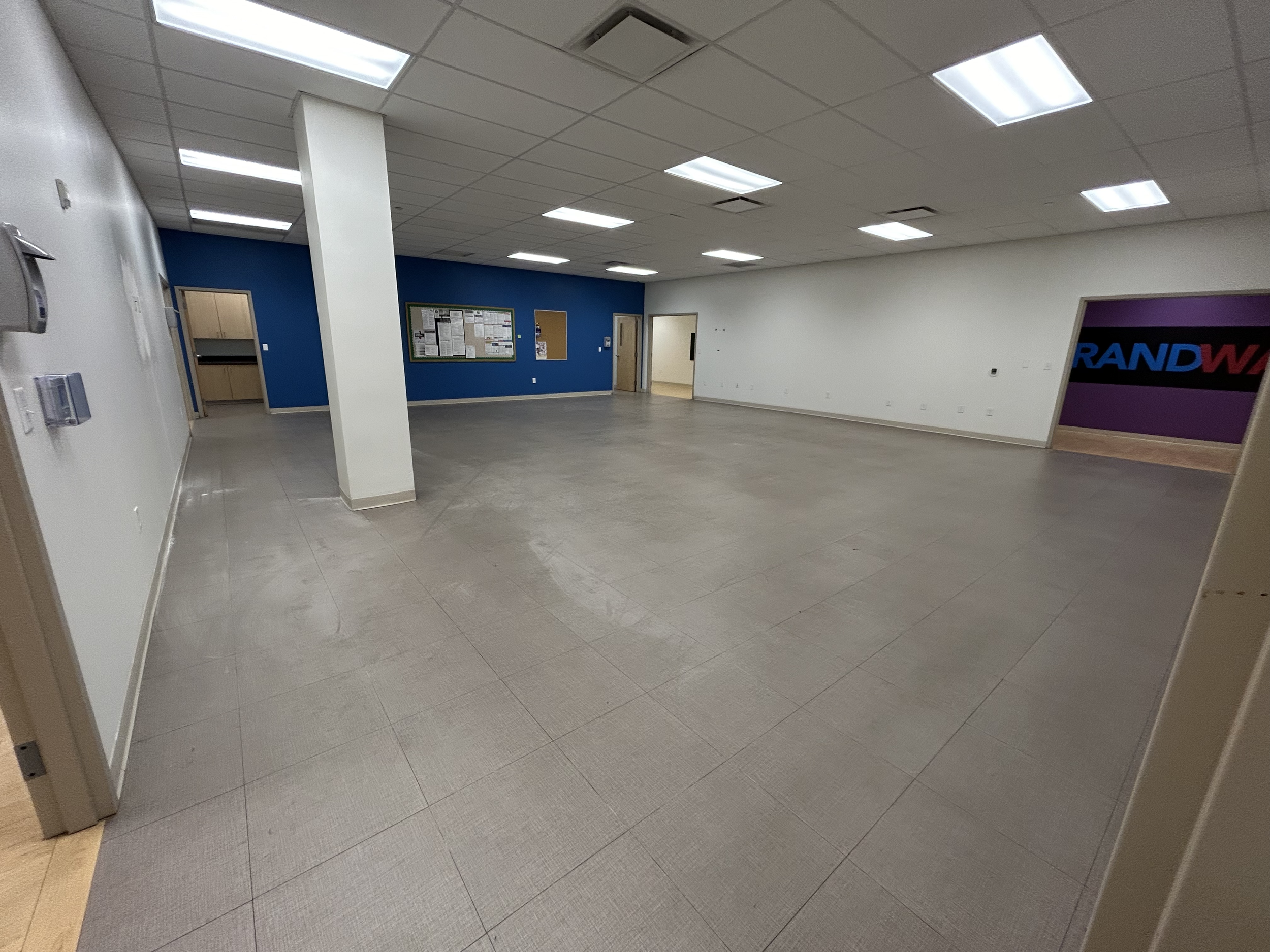 Empty room with tiled floor, white and blue walls, ceiling with fluorescent lights, bulletin board, and doorways, one with a sign that says 'RANDW'.