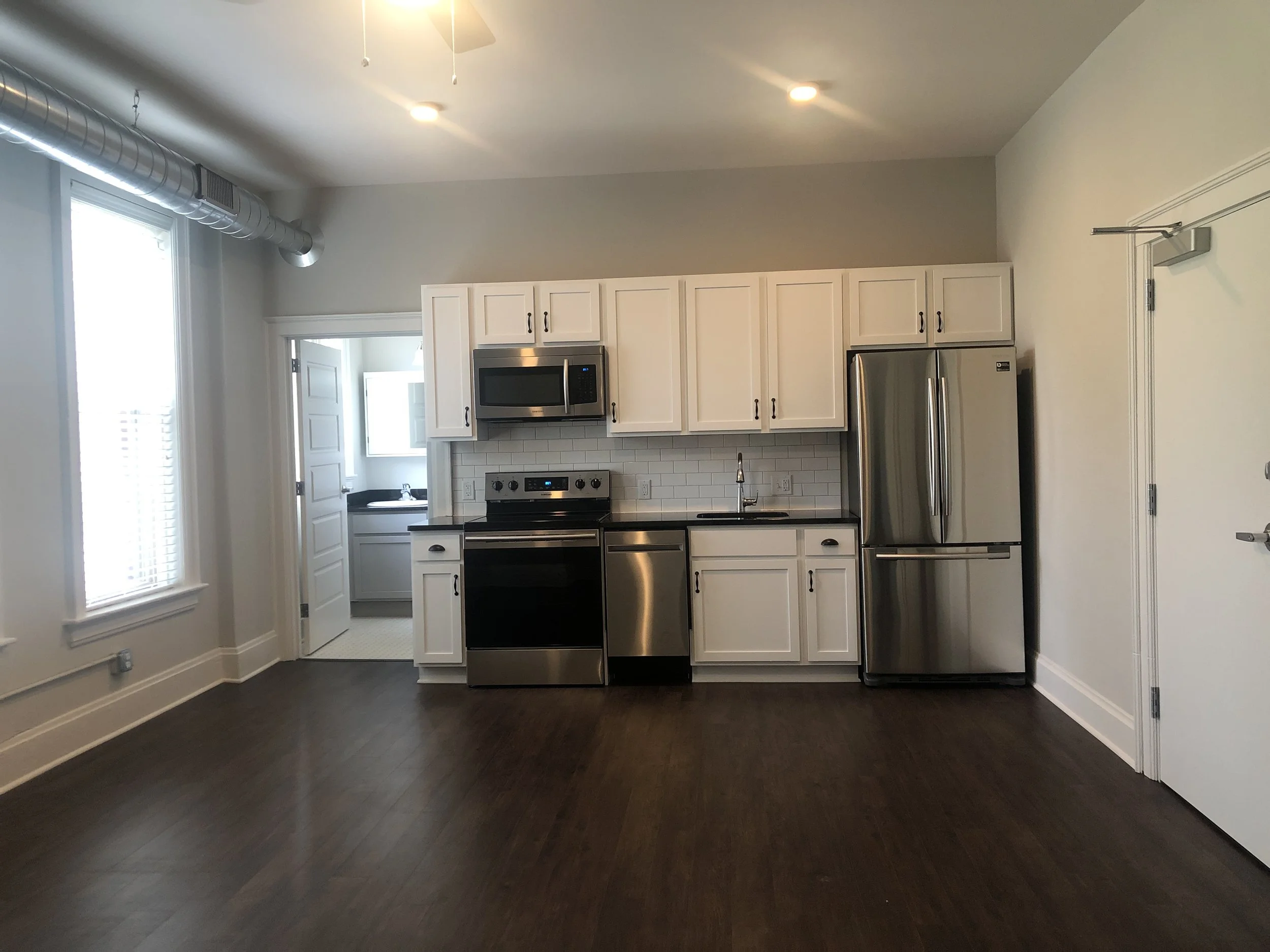 Empty modern kitchen with white cabinets, stainless steel appliances including a refrigerator, microwave, and stove, dark wood flooring, and a small adjoining laundry or utility room visible through a door.