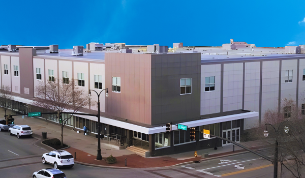 Exterior view of a modern building with a corner entrance, street signs, parked cars, and leafless trees under a clear blue sky.