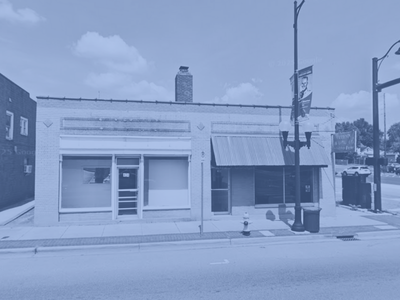 A small commercial building on a city street with large front windows, an awning, and a sidewalk with a streetlamp and trash bins.