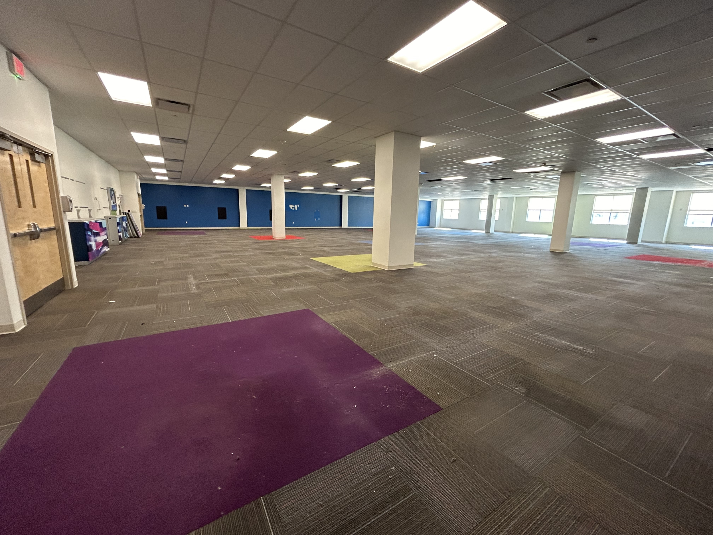 An empty, spacious indoor room with a tiled floor, white and blue walls, several large windows, ceiling lights, and colorful carpeted sections.