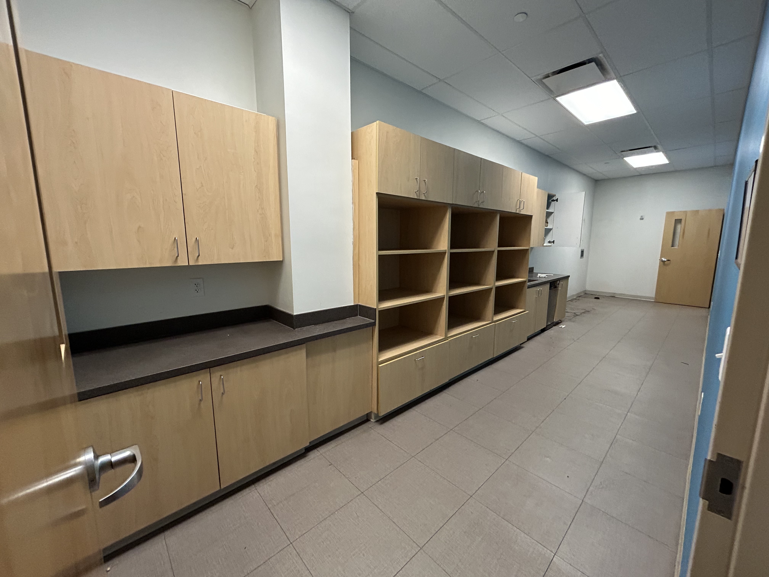 Empty room with wooden cabinets, open shelves, and a small sink area, likely a kitchen or break room in a commercial or institutional building.
