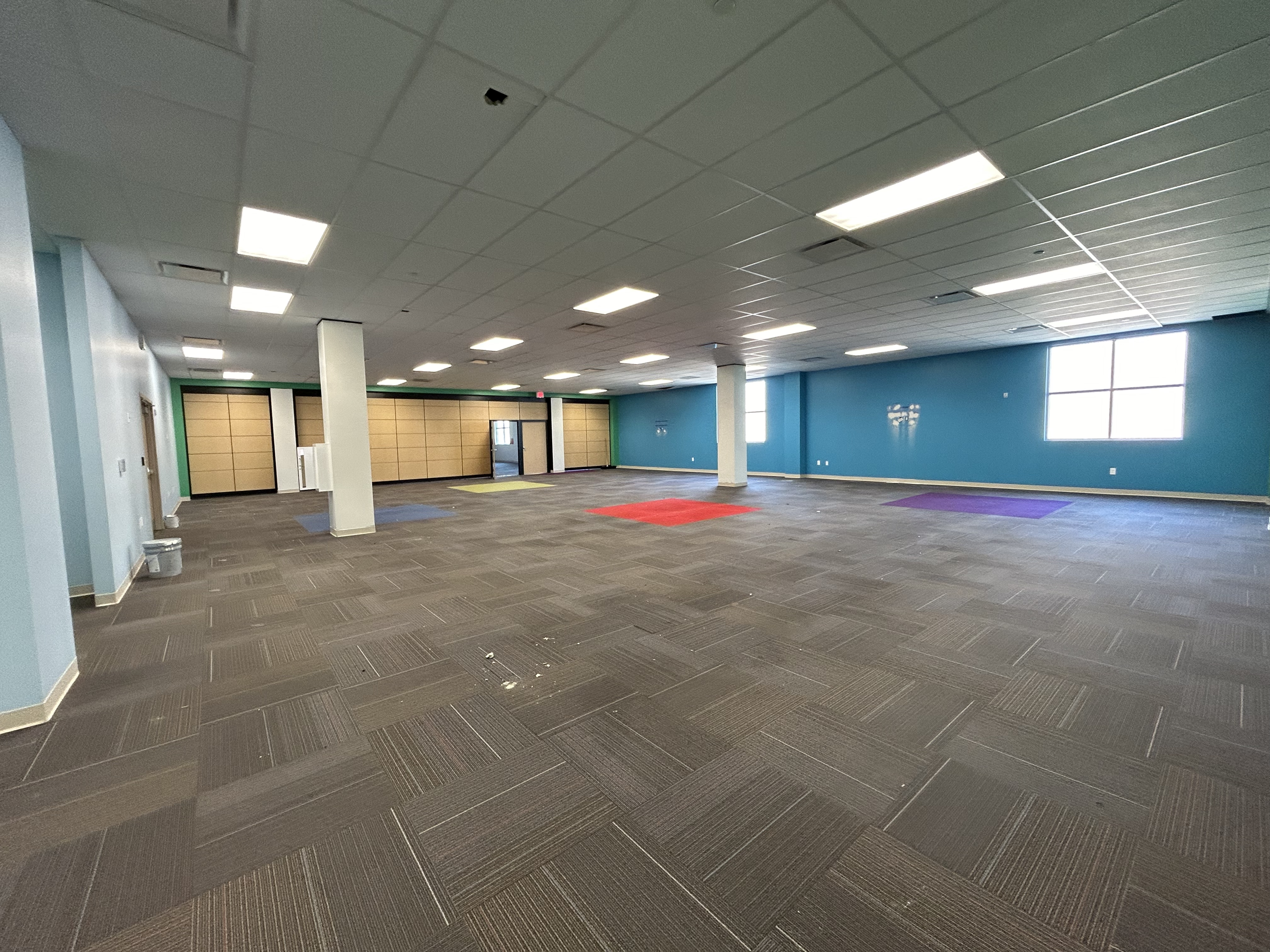 Empty multipurpose room with colorful mats, large windows, blue walls, brown carpet tiles, and a tiled ceiling with fluorescent lights.