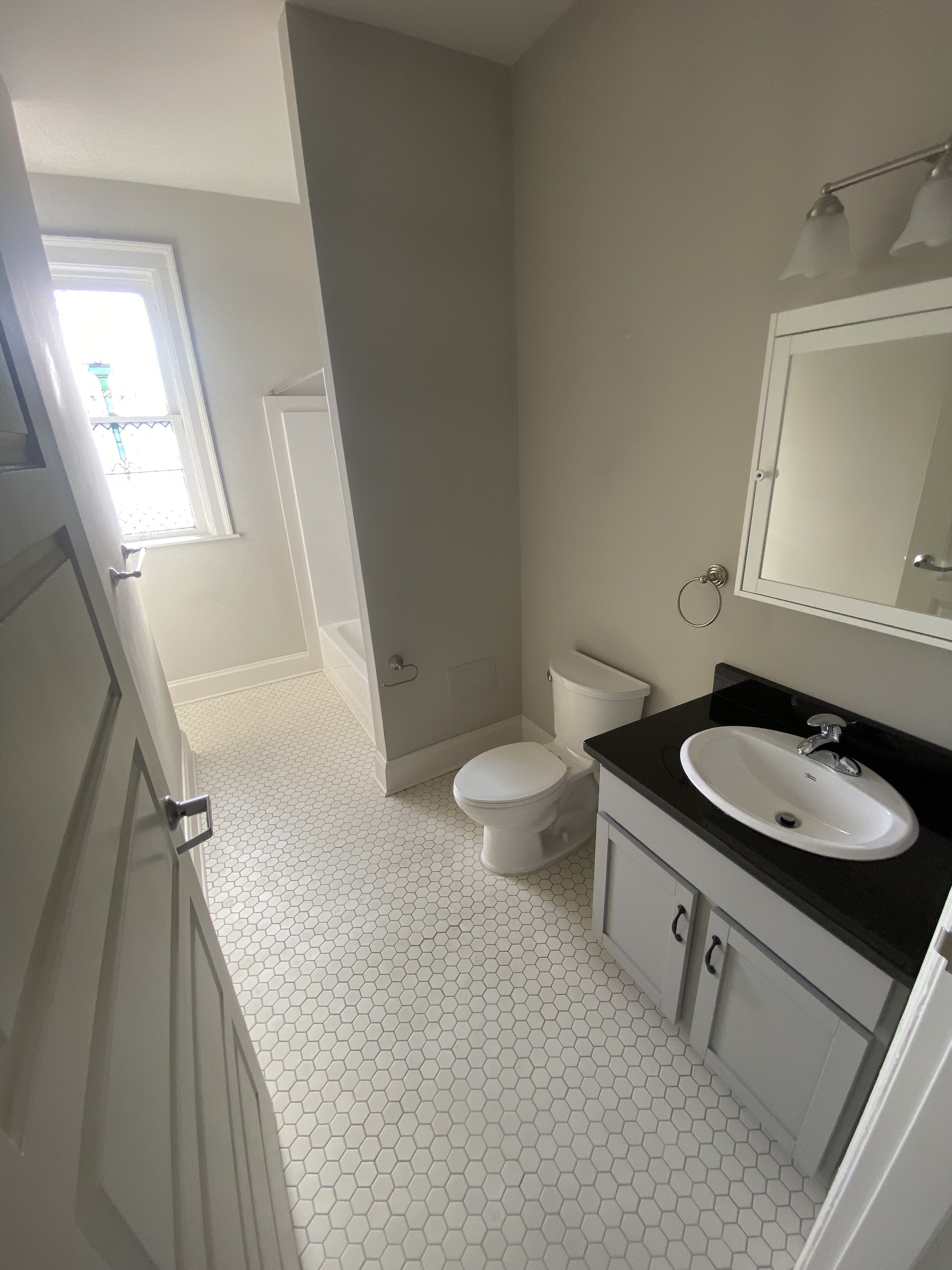 Small bathroom with white hexagon tile floor, toilet, black countertop sink with a mirror, and a window with stained glass detail in the background.