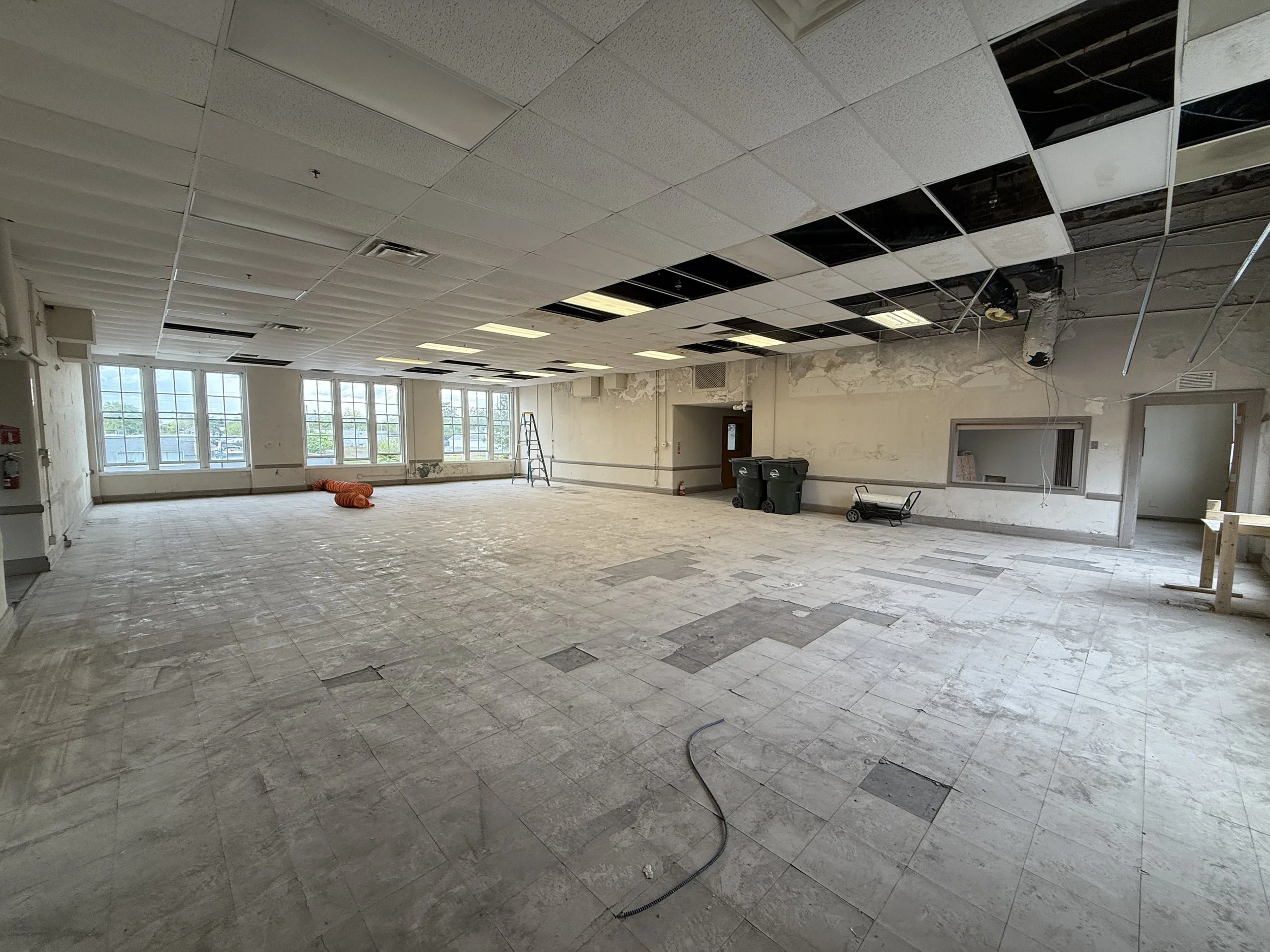 Large room under renovation with missing ceiling tiles, some ceiling panels are black. The floor is dirty with patches of missing tiles. Multiple large windows on the far wall let in natural light. Construction equipment and supplies are scattered around, including an ladder, waste bins, a cart, and a wooden frame.