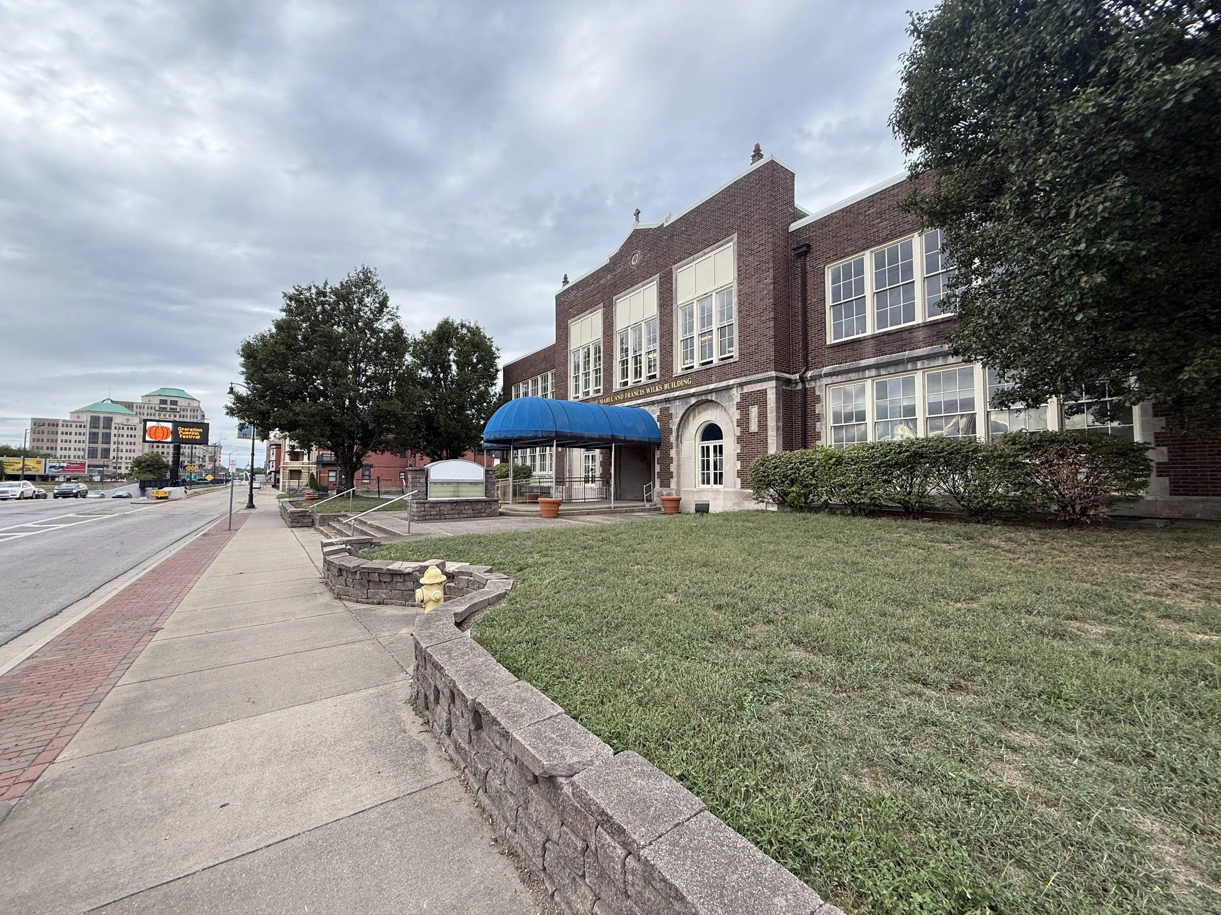A brick building with large windows and a blue awning over the entrance, next to a sidewalk and grass area, with trees and other buildings in the background under a cloudy sky.