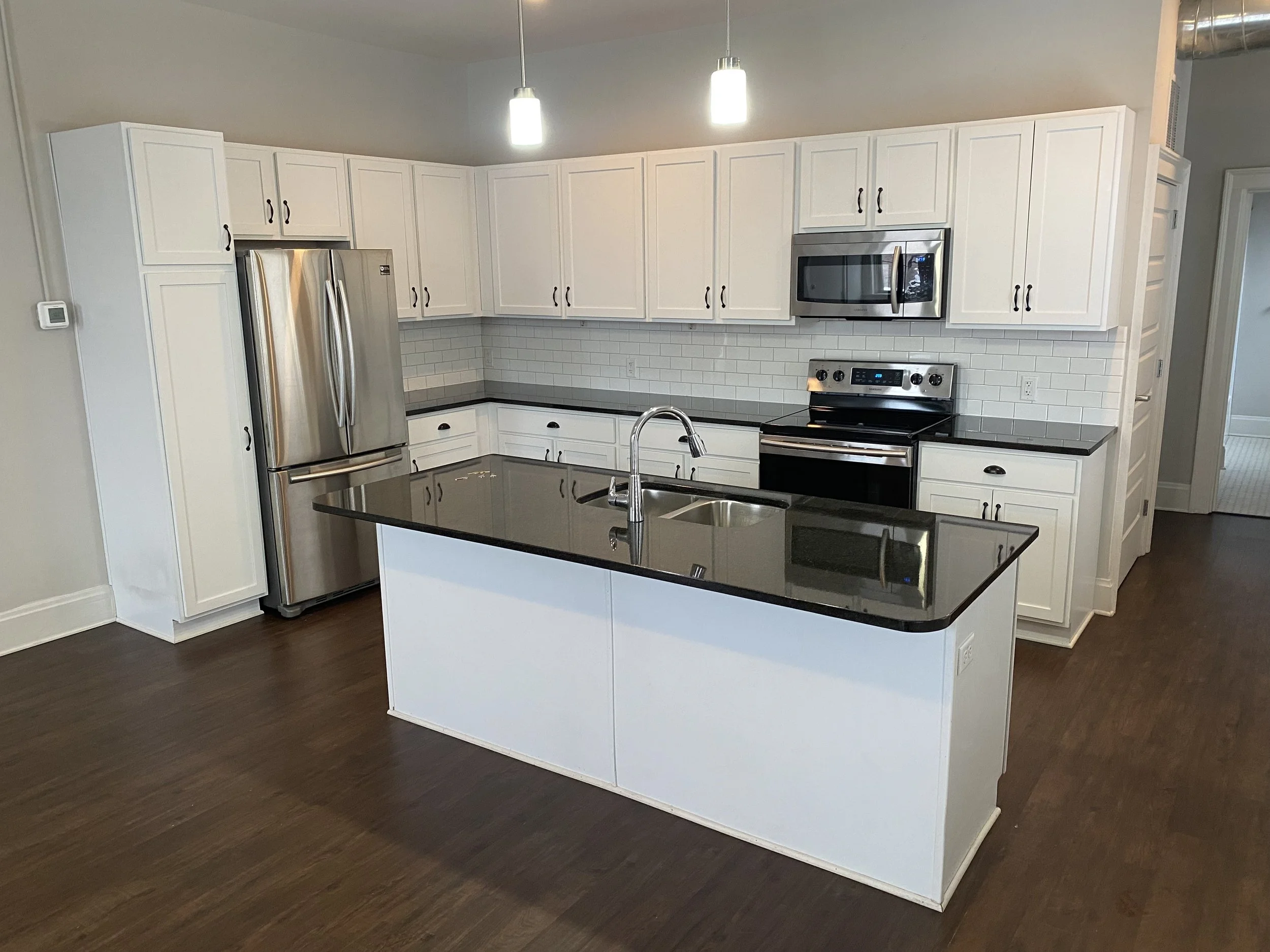 Modern kitchen with white cabinets, black countertops, stainless steel appliances including a refrigerator, microwave, and oven, a kitchen island with a black countertop, a sink with a faucet, and hardwood floors.