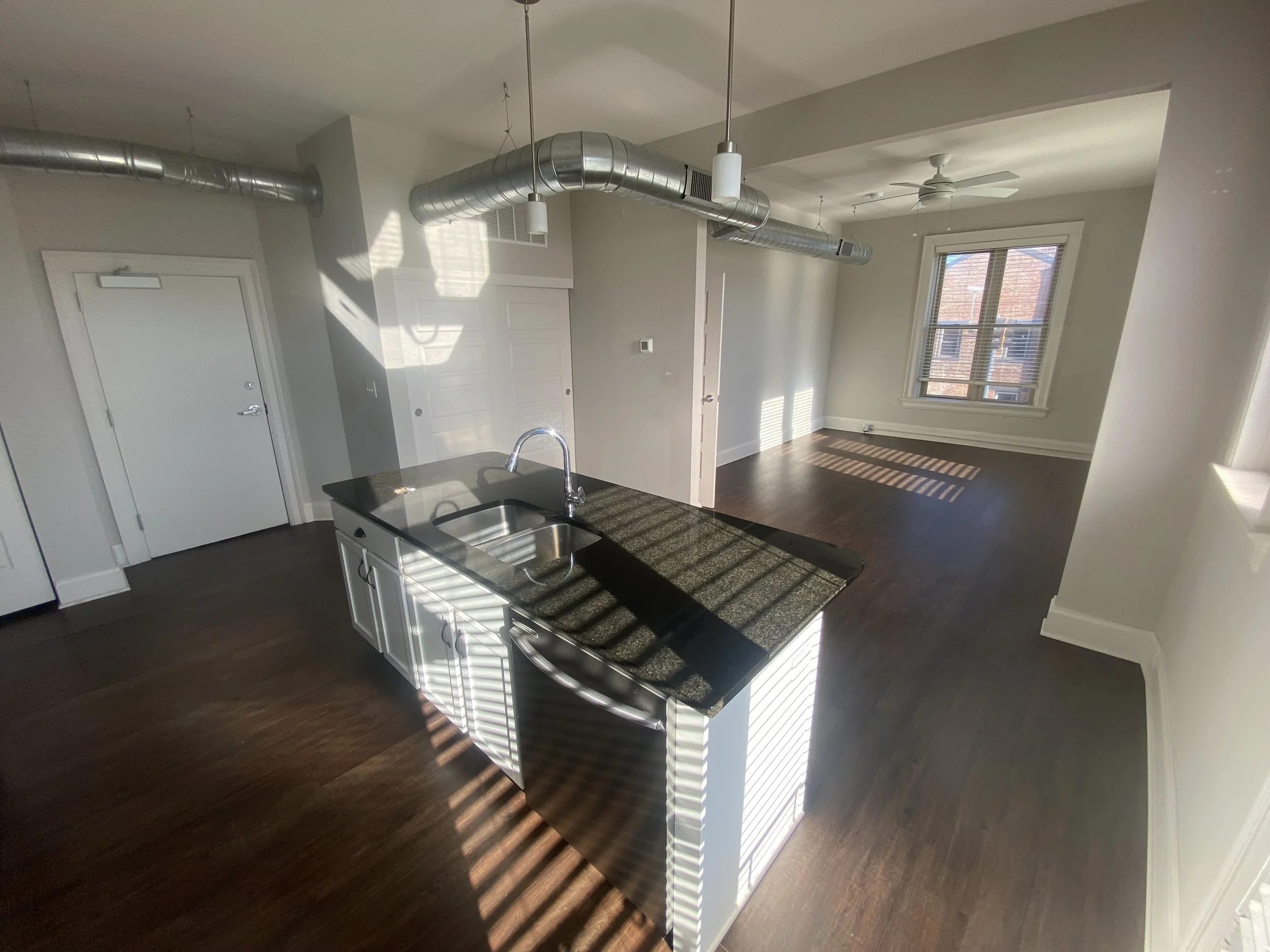 Empty apartment with an open kitchen and living area. Sunlight streams through the windows, casting shadows on the dark hardwood floor. The kitchen features a black granite countertop and a double sink, with exposed silver ductwork on the ceiling and a white ceiling fan in the living space.