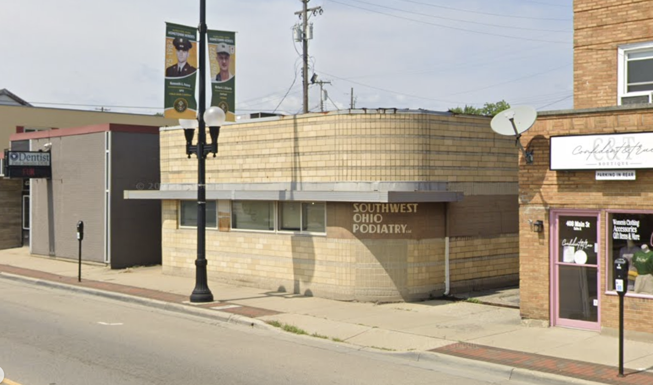 Street view of a building with a sign indicating Southwest Ohio Podiatry, next to other commercial establishments including a jewelry boutique with purple door and a cosmetic store, with banners and a streetlamp in the foreground.