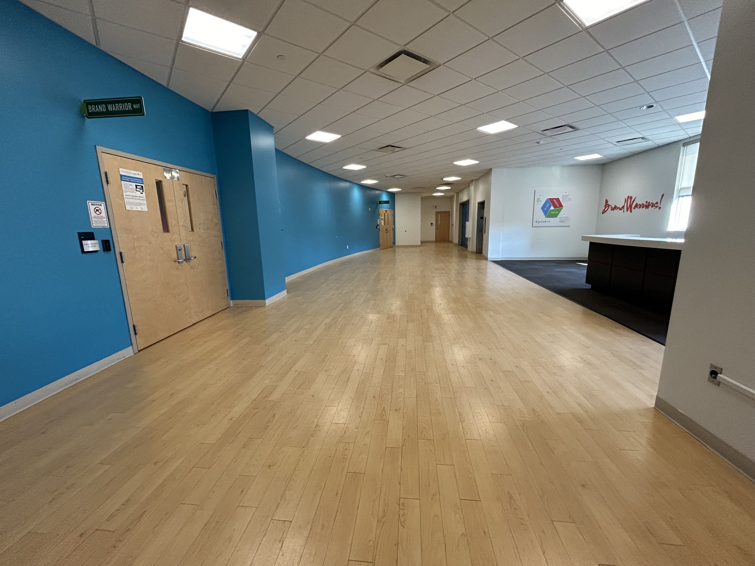 A hallway with light wood flooring, blue and white walls, ceiling lights, and a reception desk on the right. There is a sign on the blue wall that reads "Brand Warrior Way" and some double doors, along with an exit sign in the background.