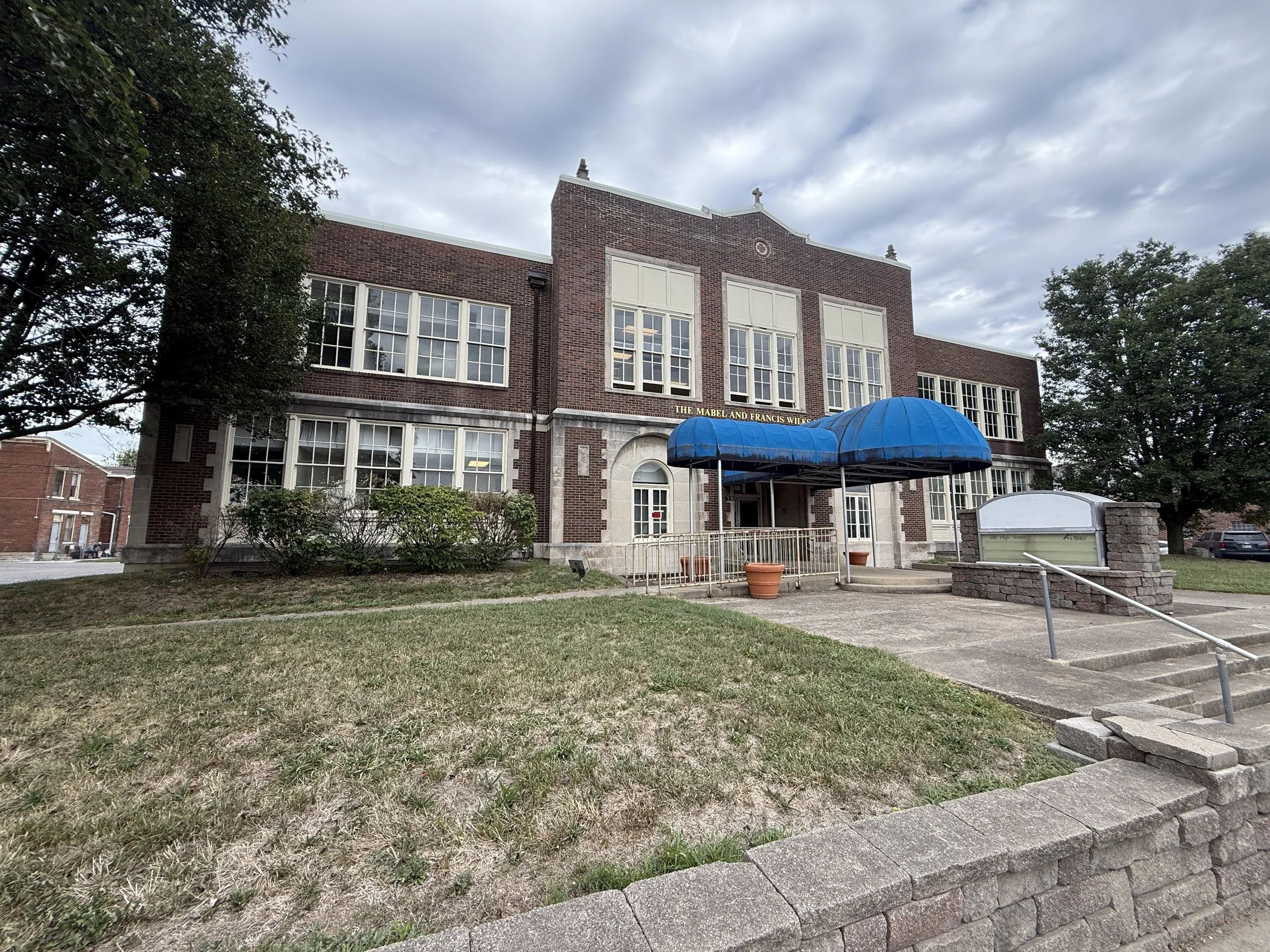Exterior of a brick school building with a blue canopy over the entrance, surrounded by grass, trees, and a concrete sidewalk, under a cloudy sky.