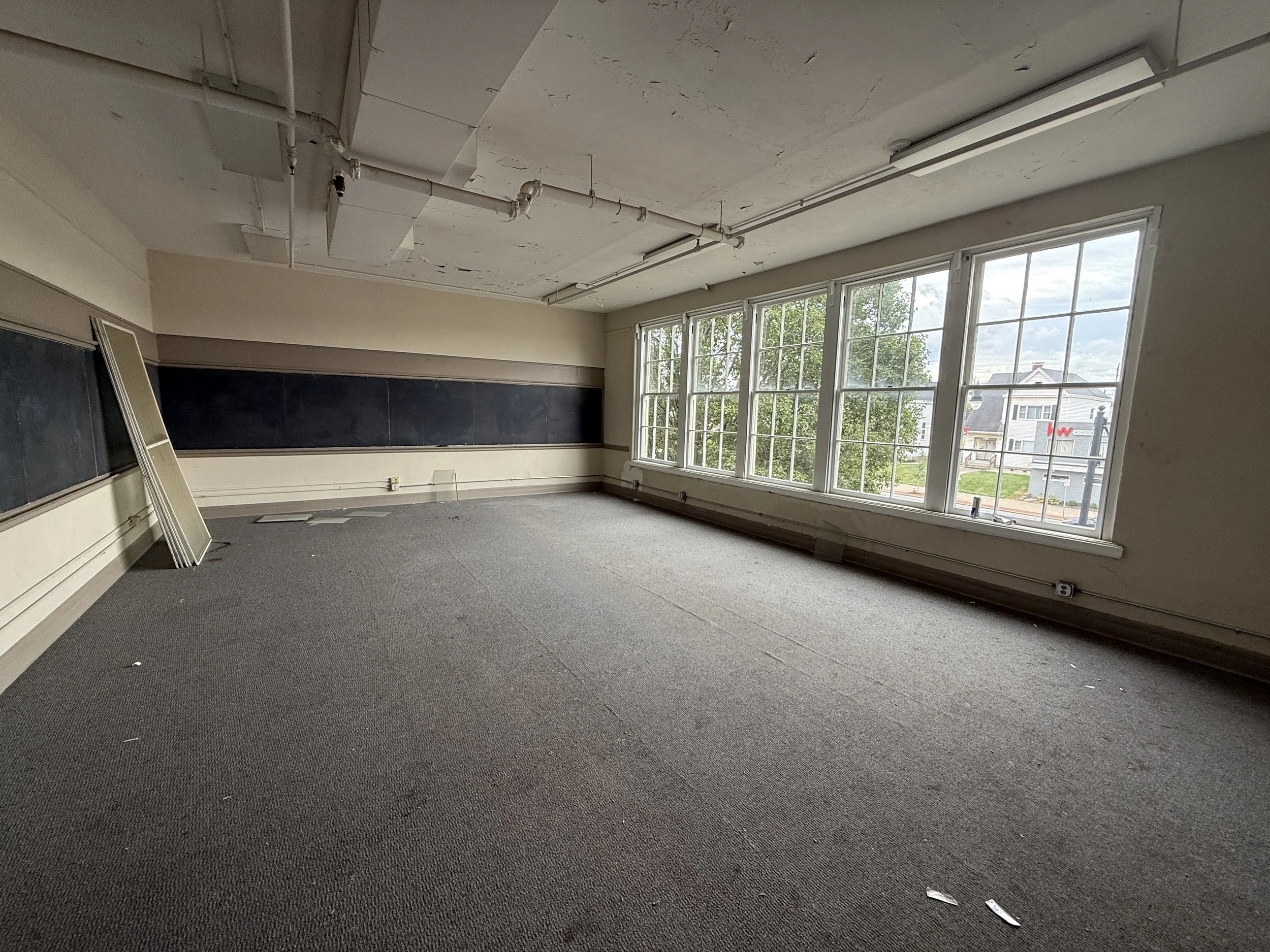 Empty classroom with blackboard on wall, large windows on one side, ceiling with exposed pipes, gray carpeted floor, leaning whiteboard or picture frame, and some debris on the floor.
