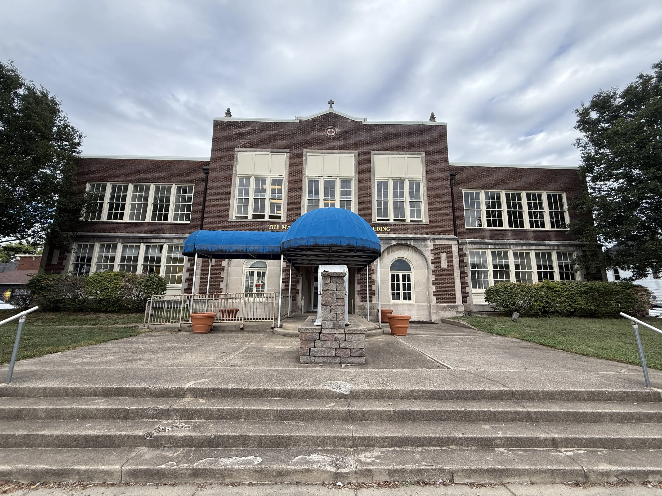 Front view of a brick school building with large windows, a blue awning over the entrance, a stone fountain in front, steps leading up, and trees on either side.