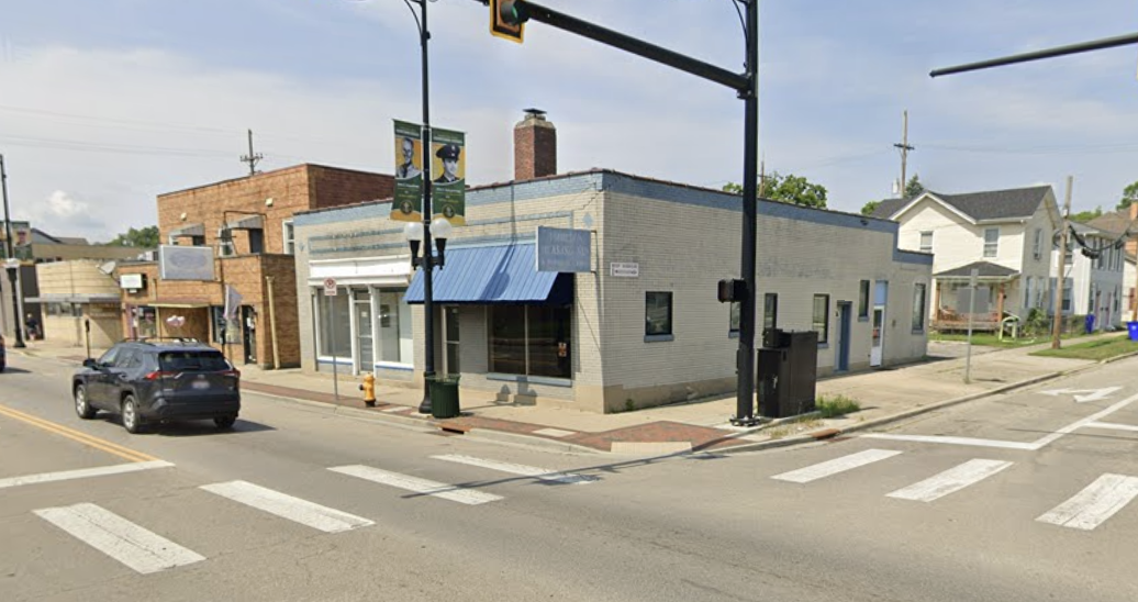 A corner of a small town street with a crosswalk, parked black SUV, and two banners hanging from a streetlight pole featuring police officers. The buildings are a mix of brick and light-colored siding, with a small sidewalk and a fire hydrant.