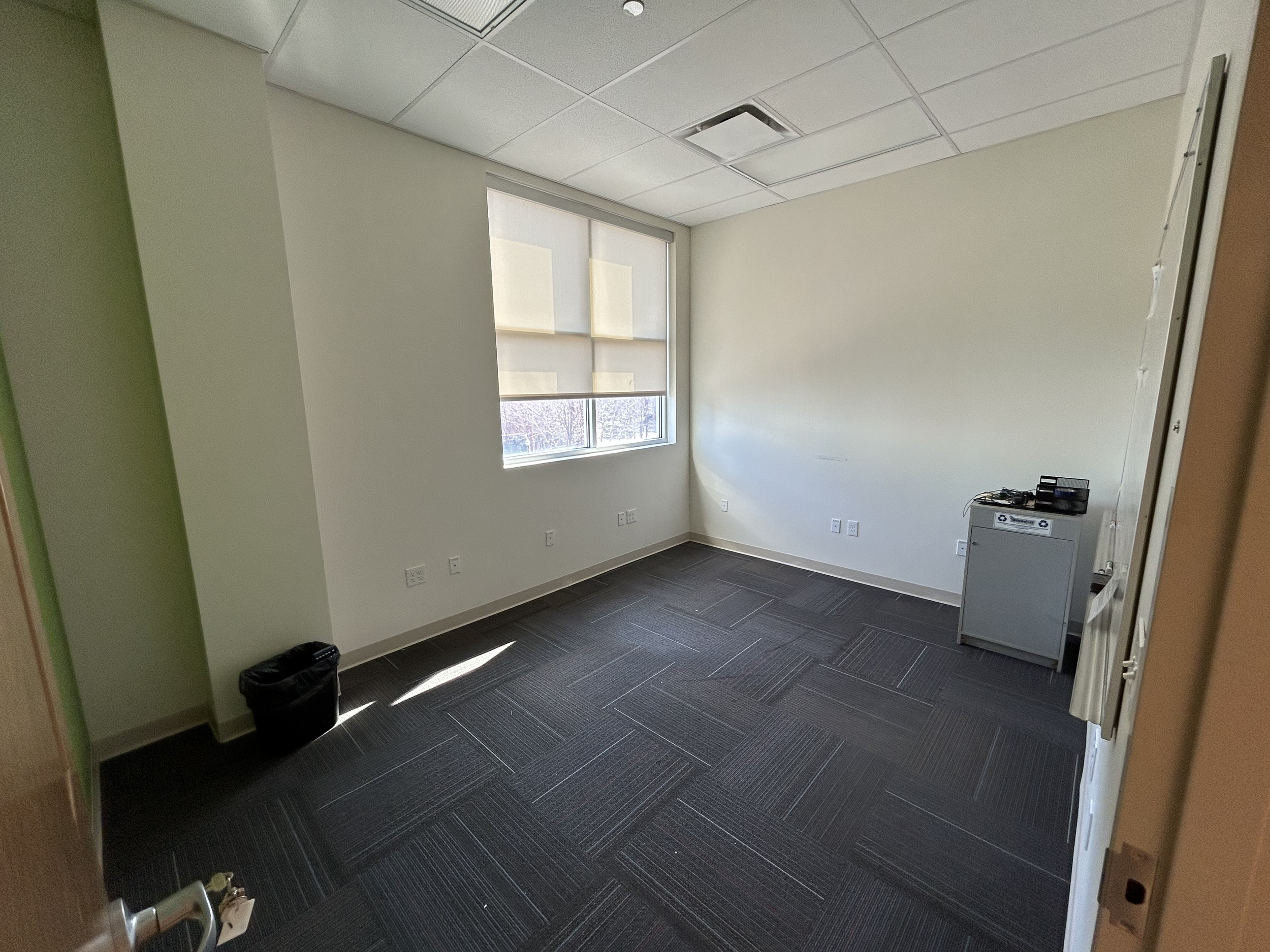 Empty office room with large window, neutral wall colors, dark carpet tiles, a small filing cabinet with electronic equipment, and a trash bin.