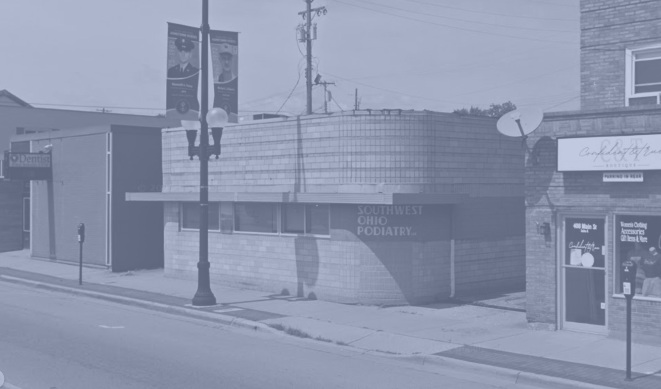 Street scene with a building housing Southwest Ohio Podiatry, a sign for a boutique, and a satellite dish, with a sidewalk, lamp post, and utility poles visible.
