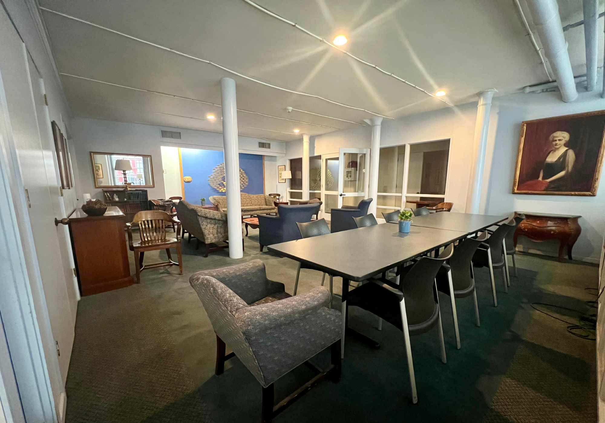 Spacious room with a large rectangular table surrounded by black chairs, a vintage armchair, a portrait painting on the wall, and various sofas and armchairs in the background.