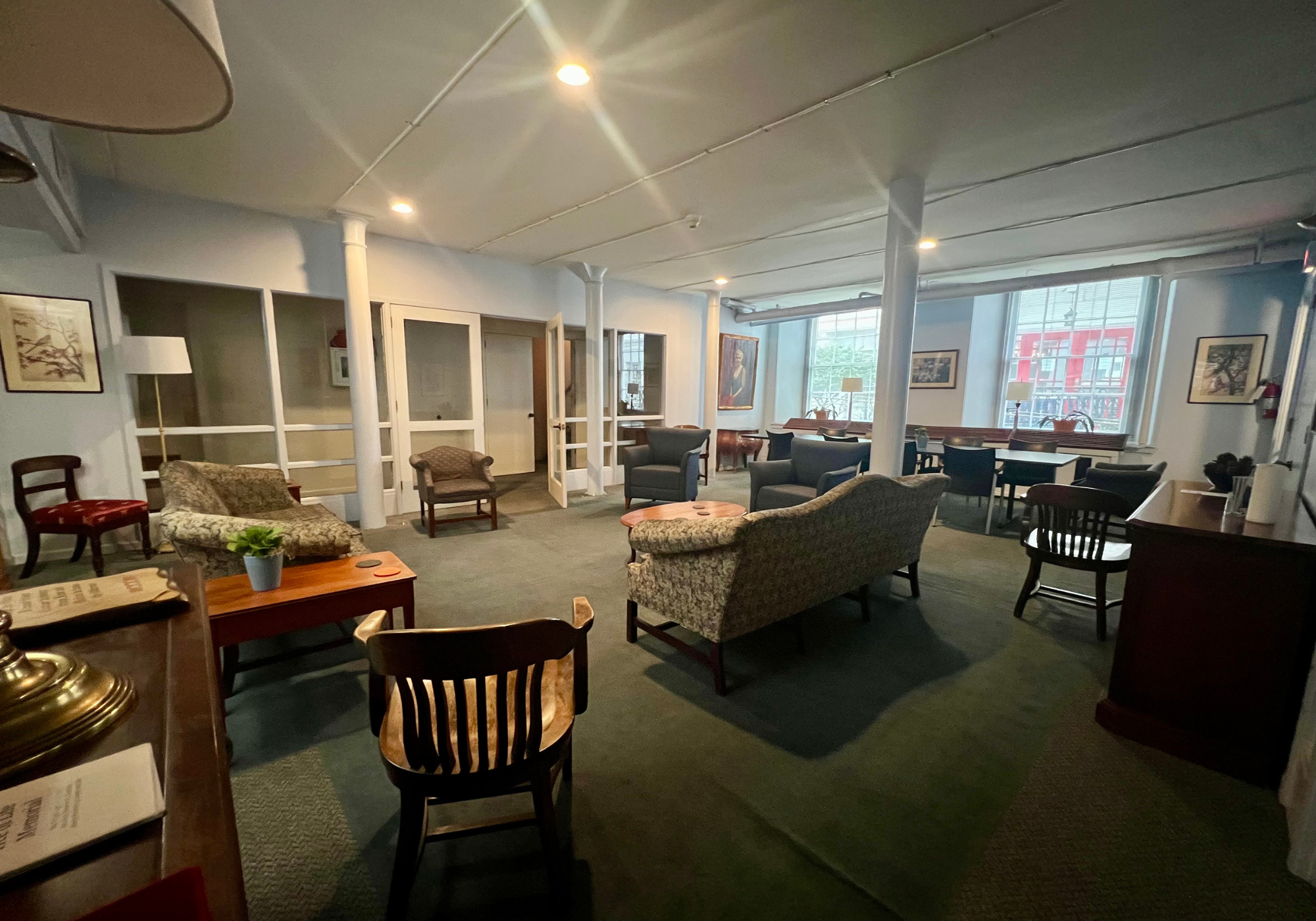 A spacious lounge area with various chairs and sofas, potted plant on a small table, large windows letting in natural light, framed artwork on the walls, and a mix of vintage and modern furniture.