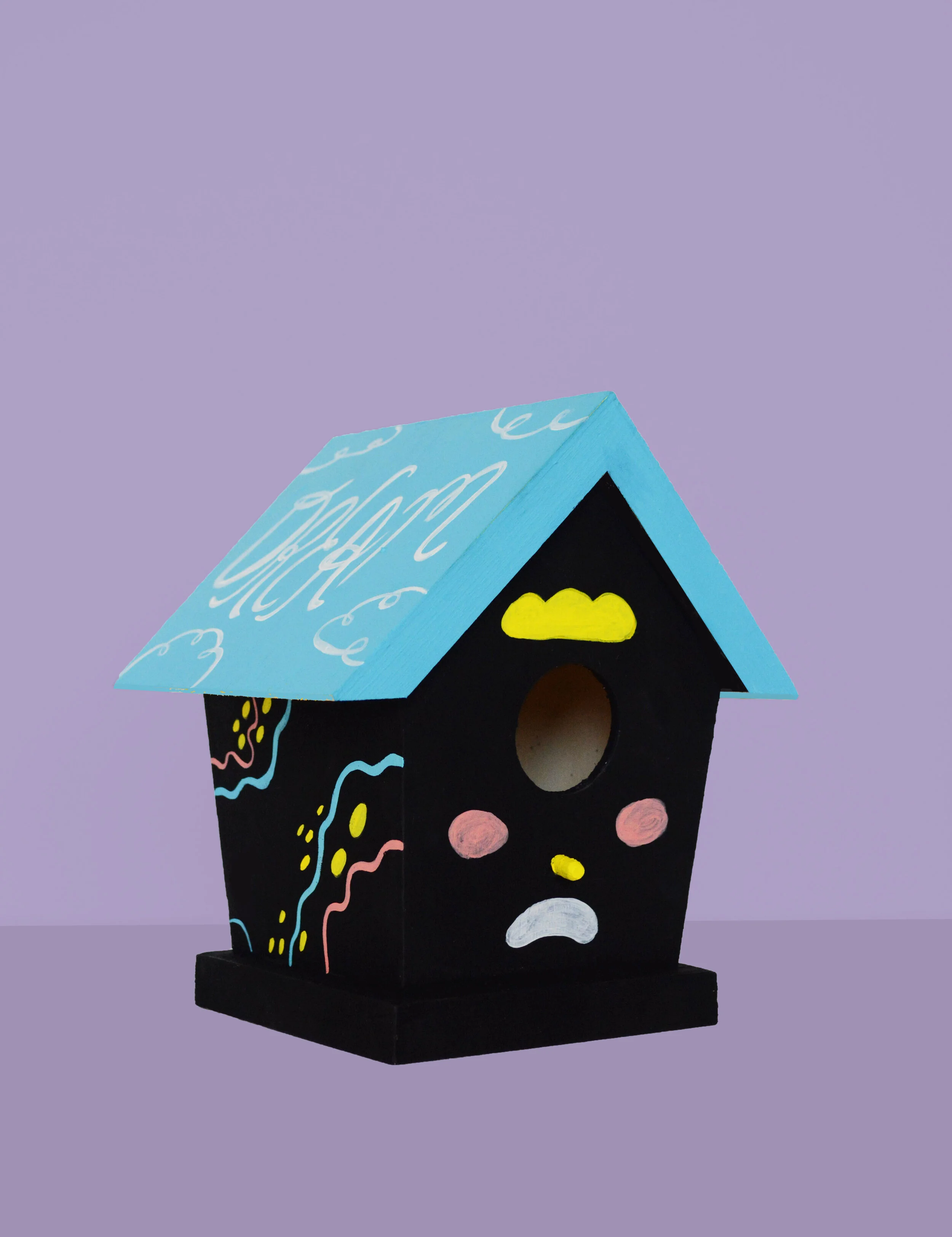 A colorful birdhouse painted black with a blue roof featuring white cursive writing, decorated with pink, yellow, white, and blue designs, and displaying a sad face with a yellow cloud above the entrance.