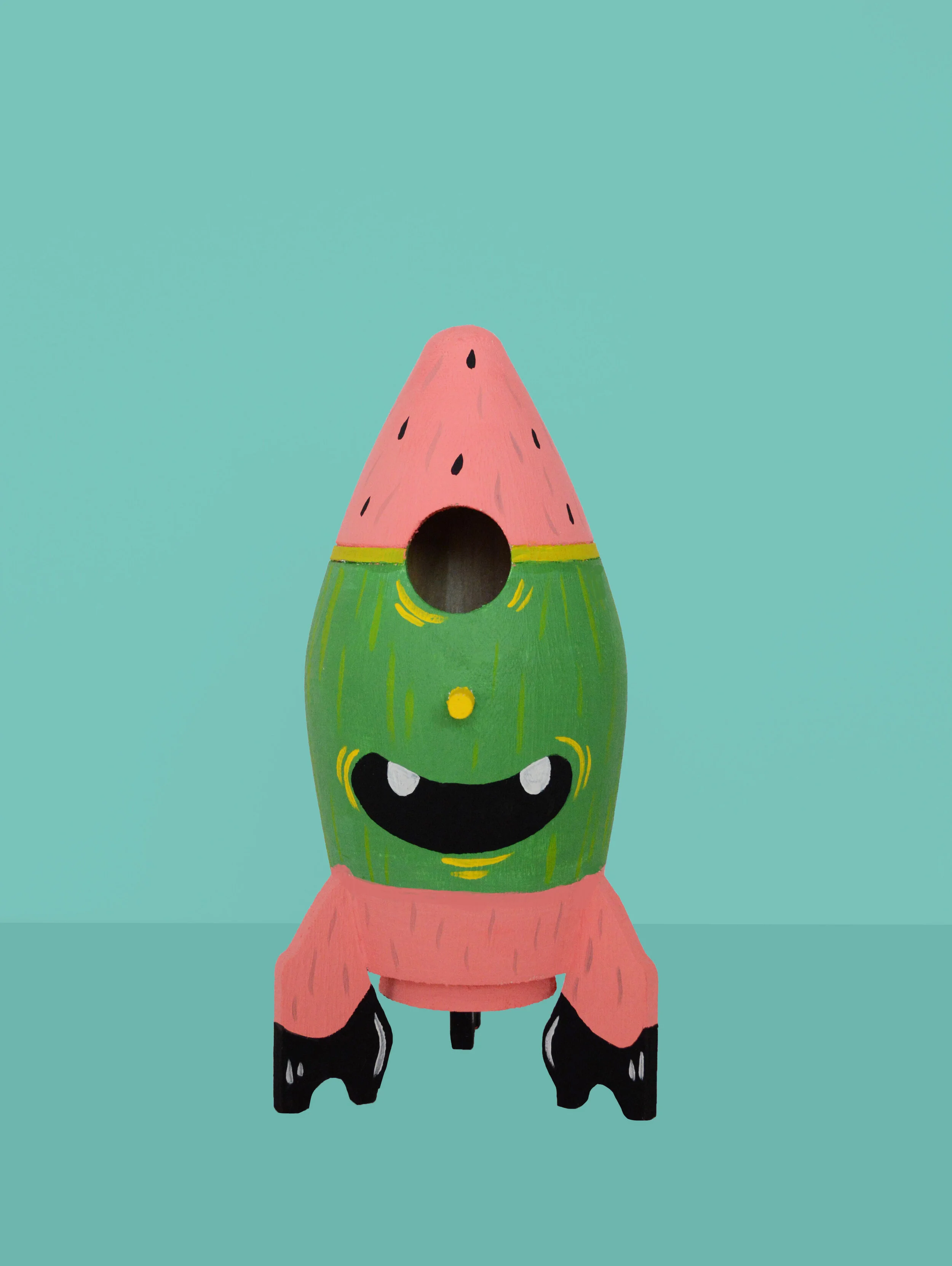 Colorful rocket shaped object with watermelon design, smiling face, and pink fins, on a teal background.