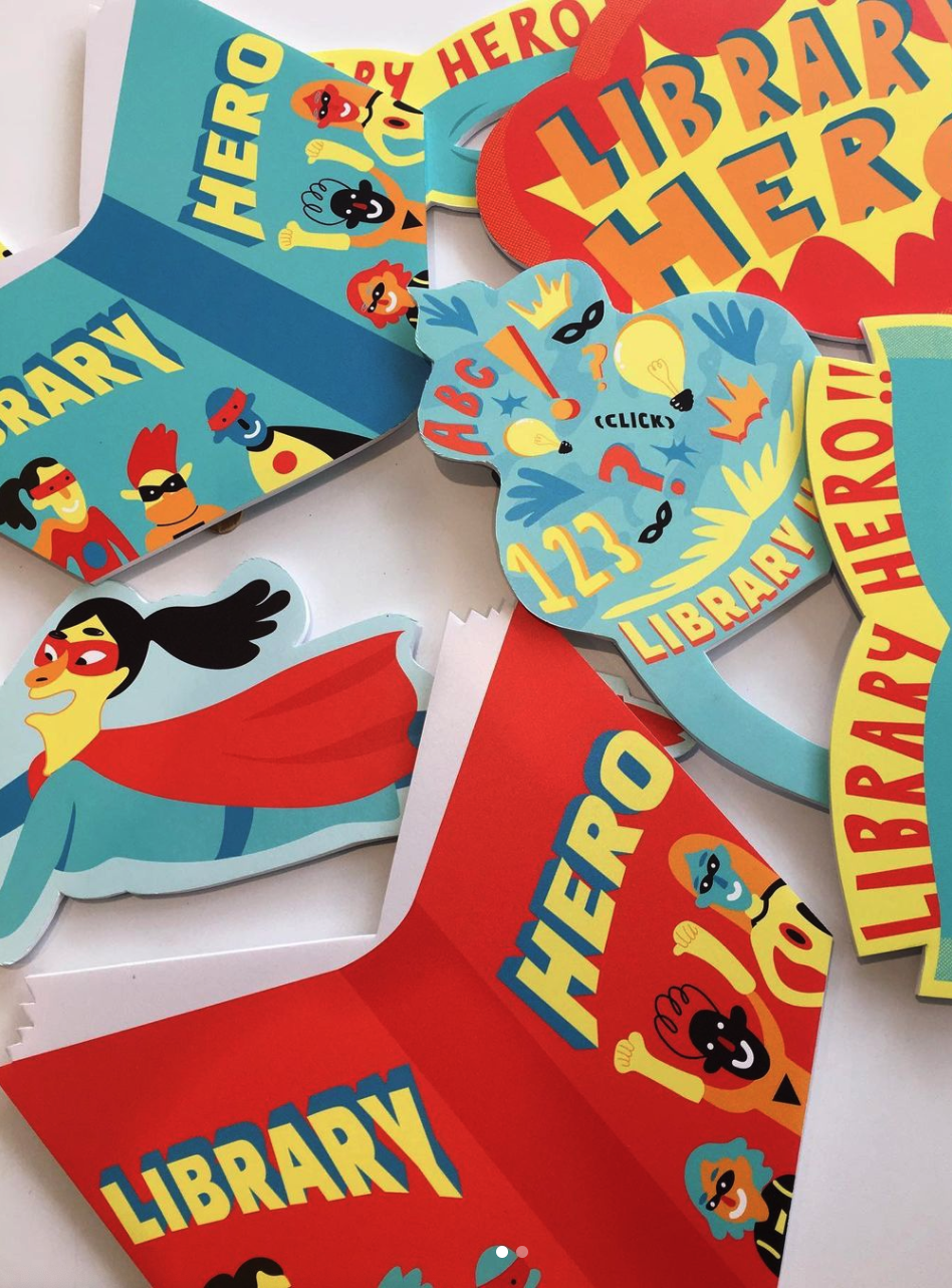 Colorful superhero-themed paper cutouts and signs for a children's library event. The signs feature words like 'LIBRARY' and 'HERO,' cartoon superhero characters with masks, capes, and cheerful expressions, and decorative elements like stars and excl