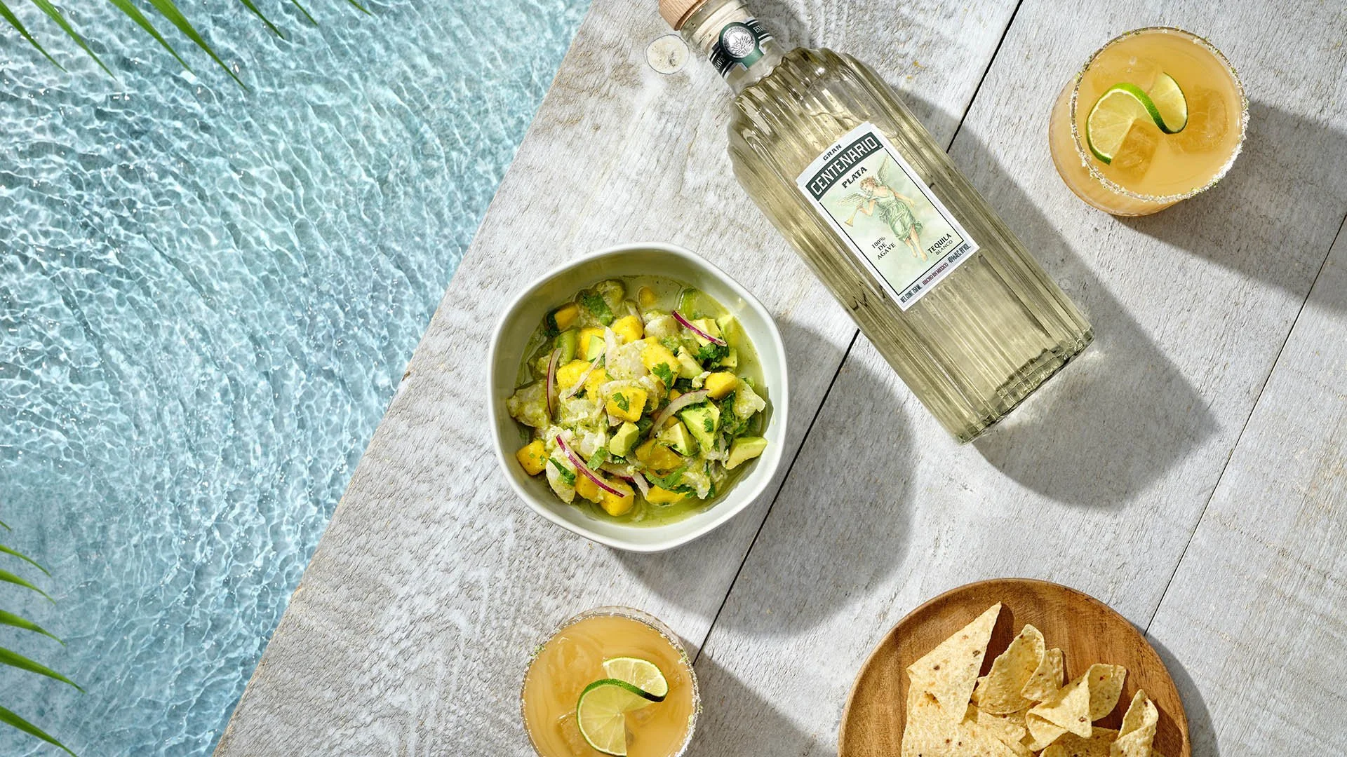 Overhead view of a poolside setup with a bowl of guacamole, a bottle of tequila, two drinks with lemon and lime slices, and a wooden plate of tortilla chips on a white wooden surface.