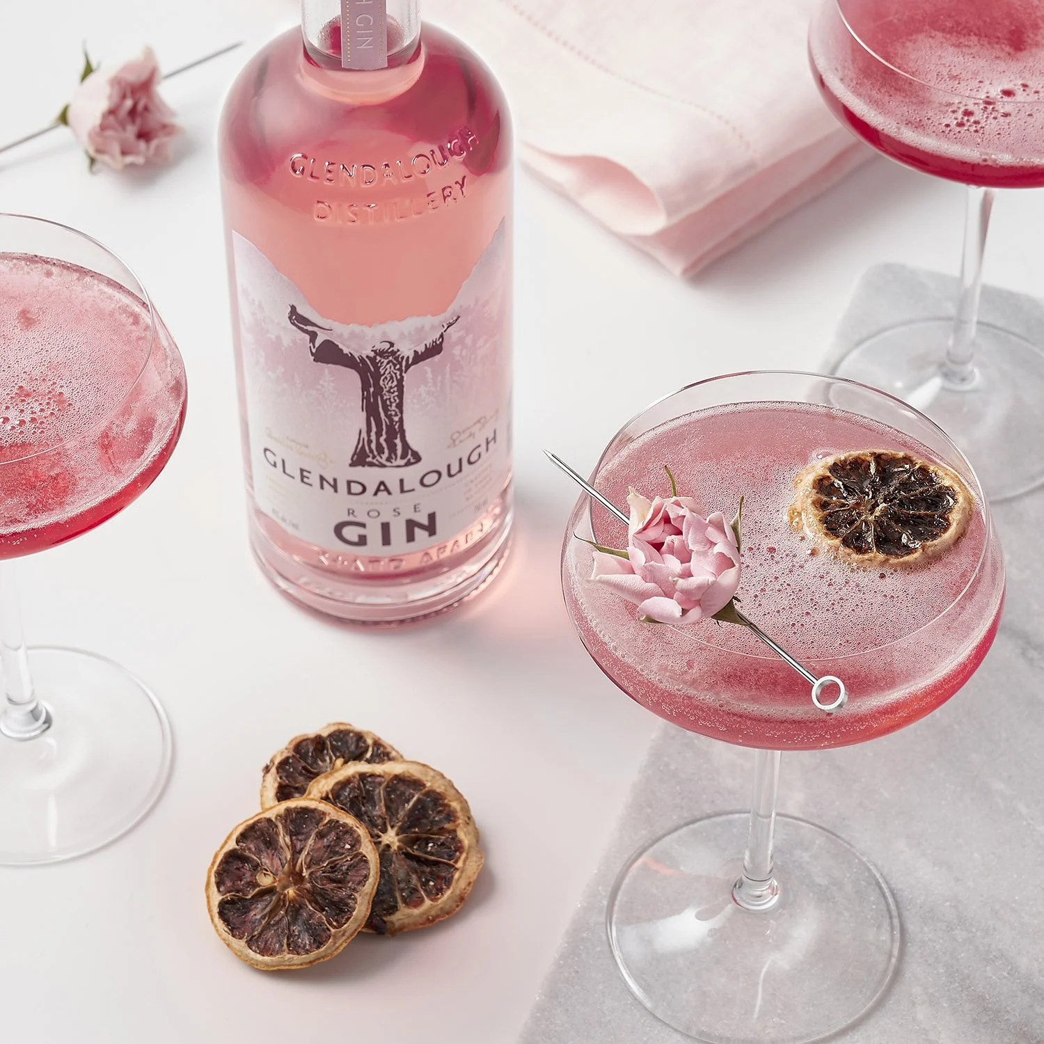 A bottle of Glendalough Rose Gin, two pink cocktails garnished with dried citrus slices and flowers, and dried citrus slices on a white surface.