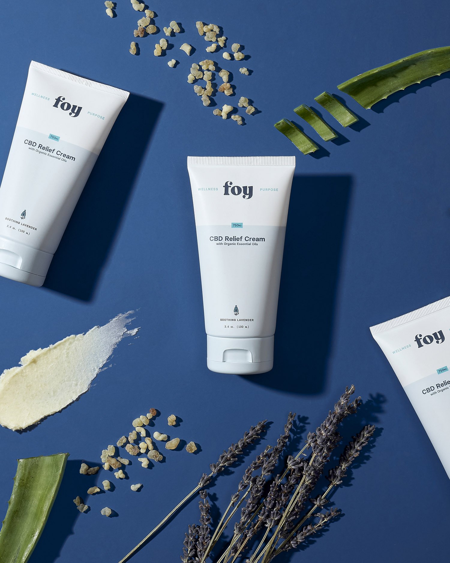 Three tubes of Foy CBD Relief Cream with lavender and herbal elements on a blue background.