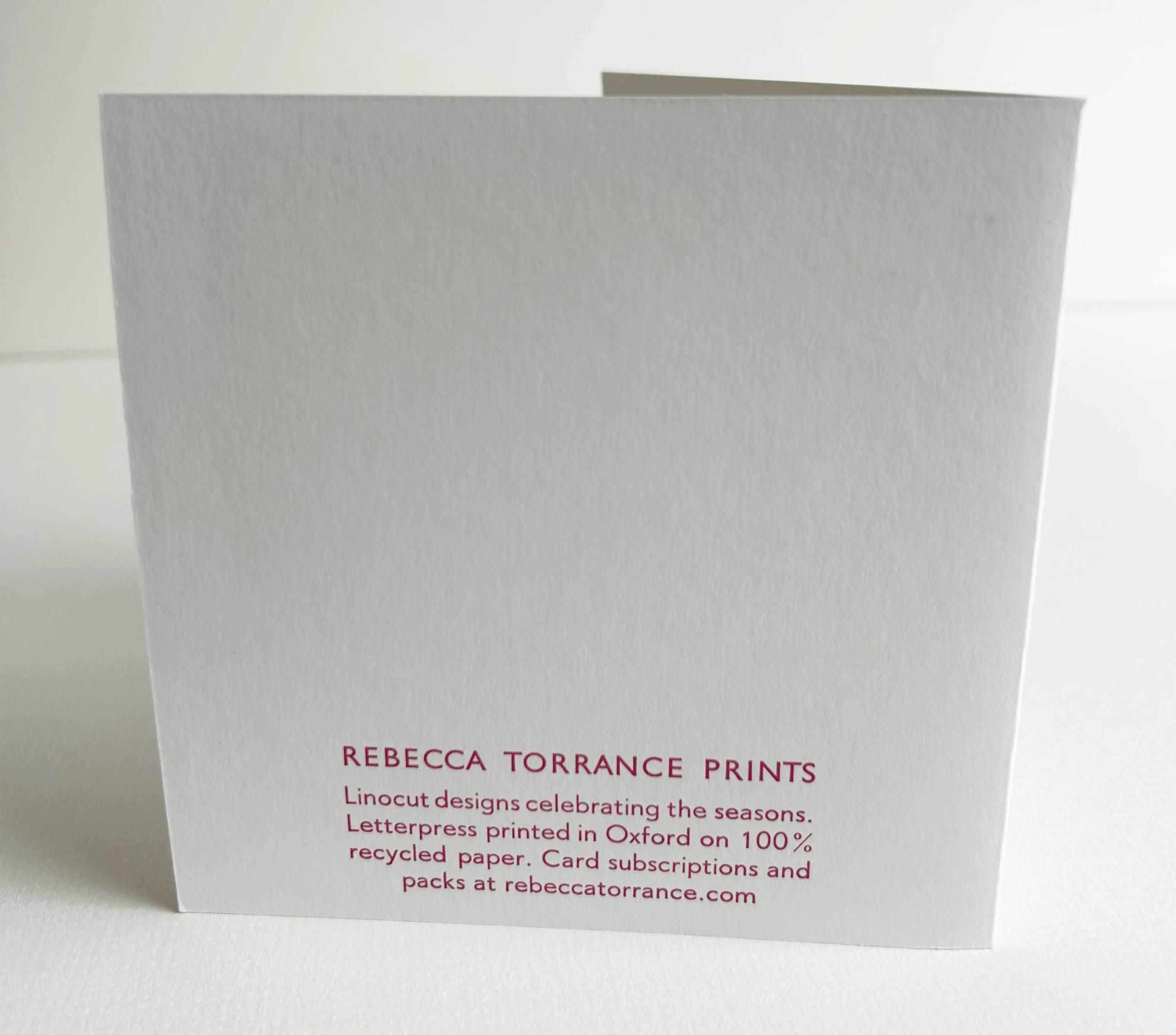 White paper card with pink text that reads 'Rebecca Torrance Prints' and details about celebrating the seasons with letterpress designs on recycled paper, available at rebeccatorrance.com.