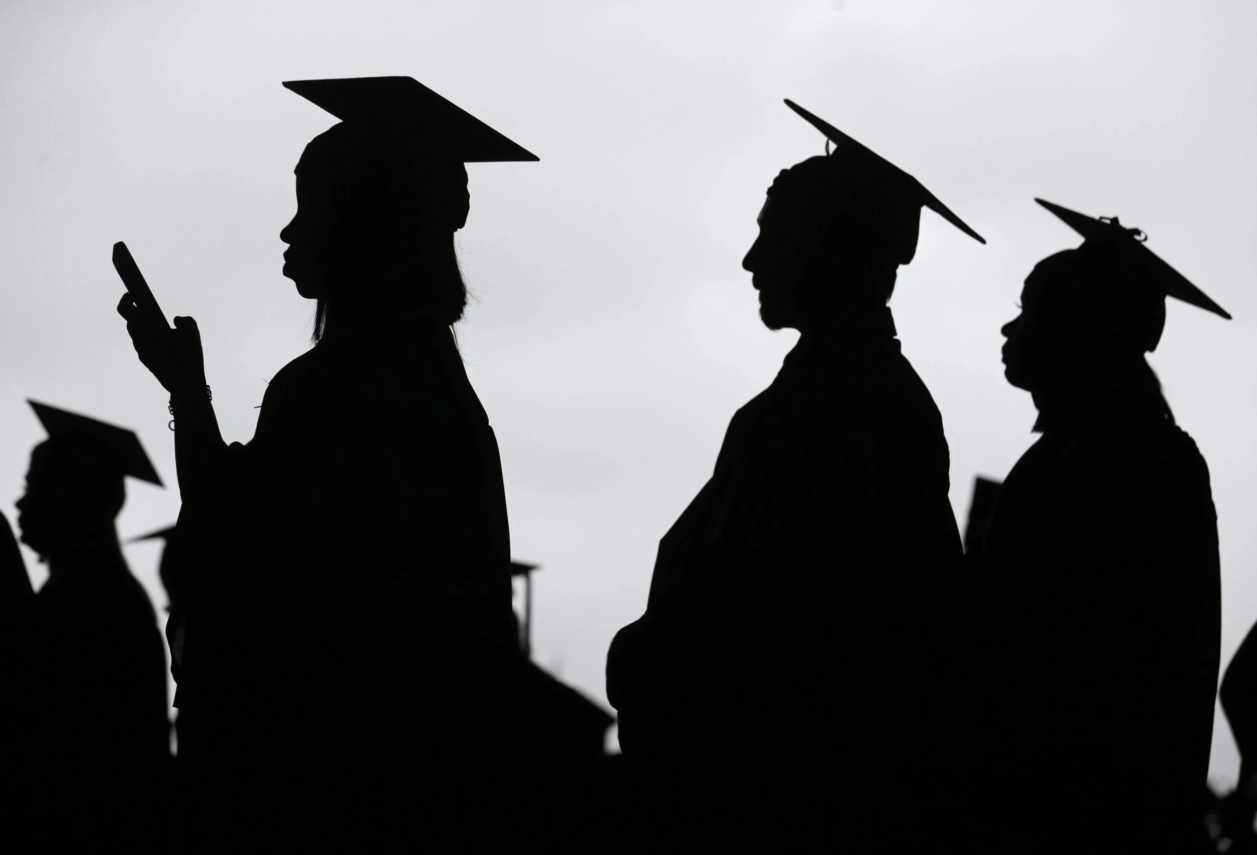 Silhouettes of people wearing graduation caps, with one person looking at a mobile device, against a gray sky.