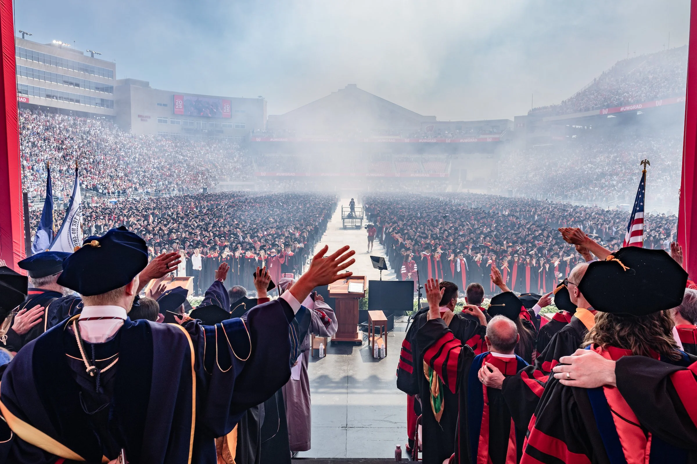 Large graduation ceremony with graduates in caps and gowns standing on stage and audience clapping, taken from inside a building with open doors showing the outdoor stadium filled with attendees and smoke or mist.