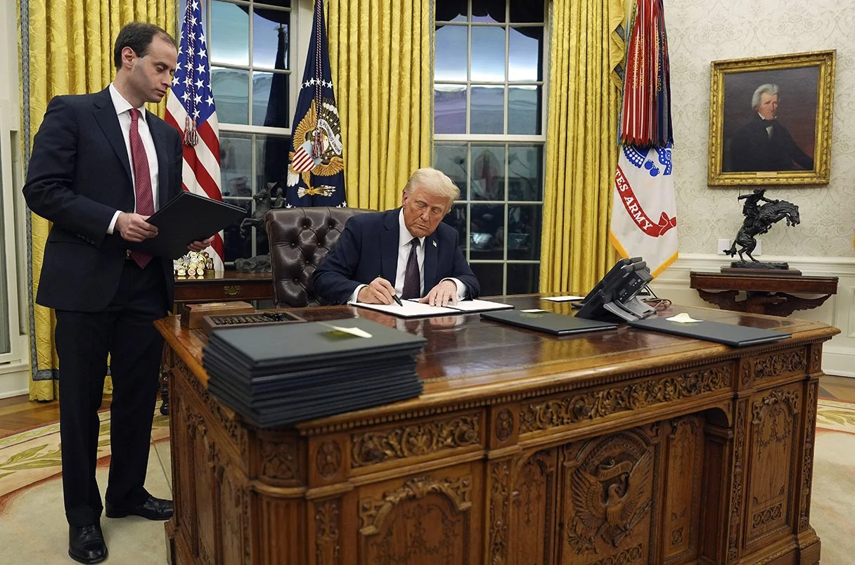 Donald Trump sitting at the Oval Office desk, signing a document with a black pen, while a man in a suit and red tie stands beside him holding a folder. The desk has a large stack of folders on it. The background shows yellow curtains, windows, flags, a framed portrait, and a decorative sculpture.