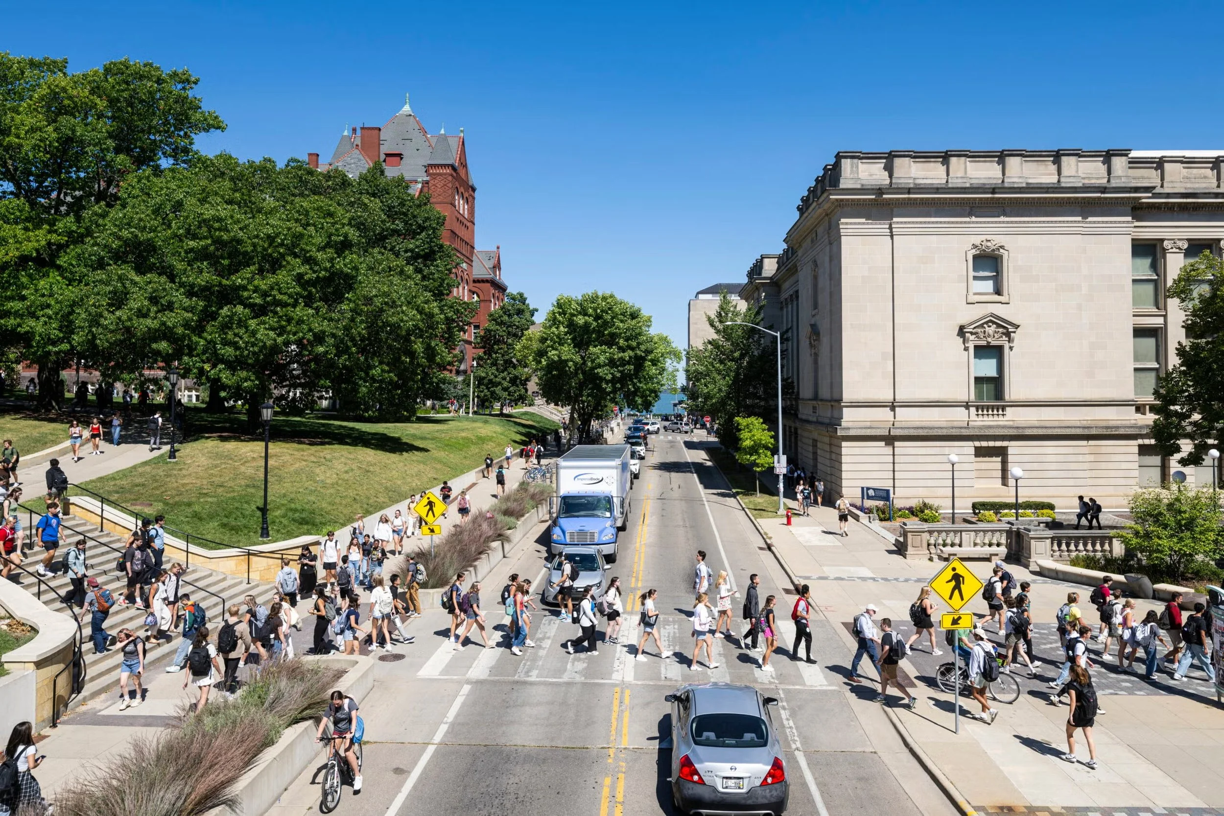 City street scene with large crowd of pedestrians crossing at crosswalk, cars on the road, historic buildings on the right, green trees on the left, and bright blue sky overhead.