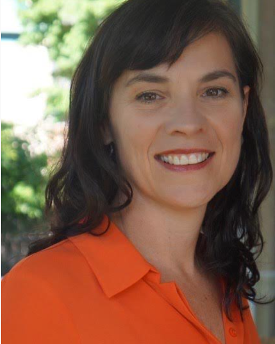 Close-up of a woman smiling, wearing an orange collared shirt, with dark wavy hair and a blurred green outdoor background.