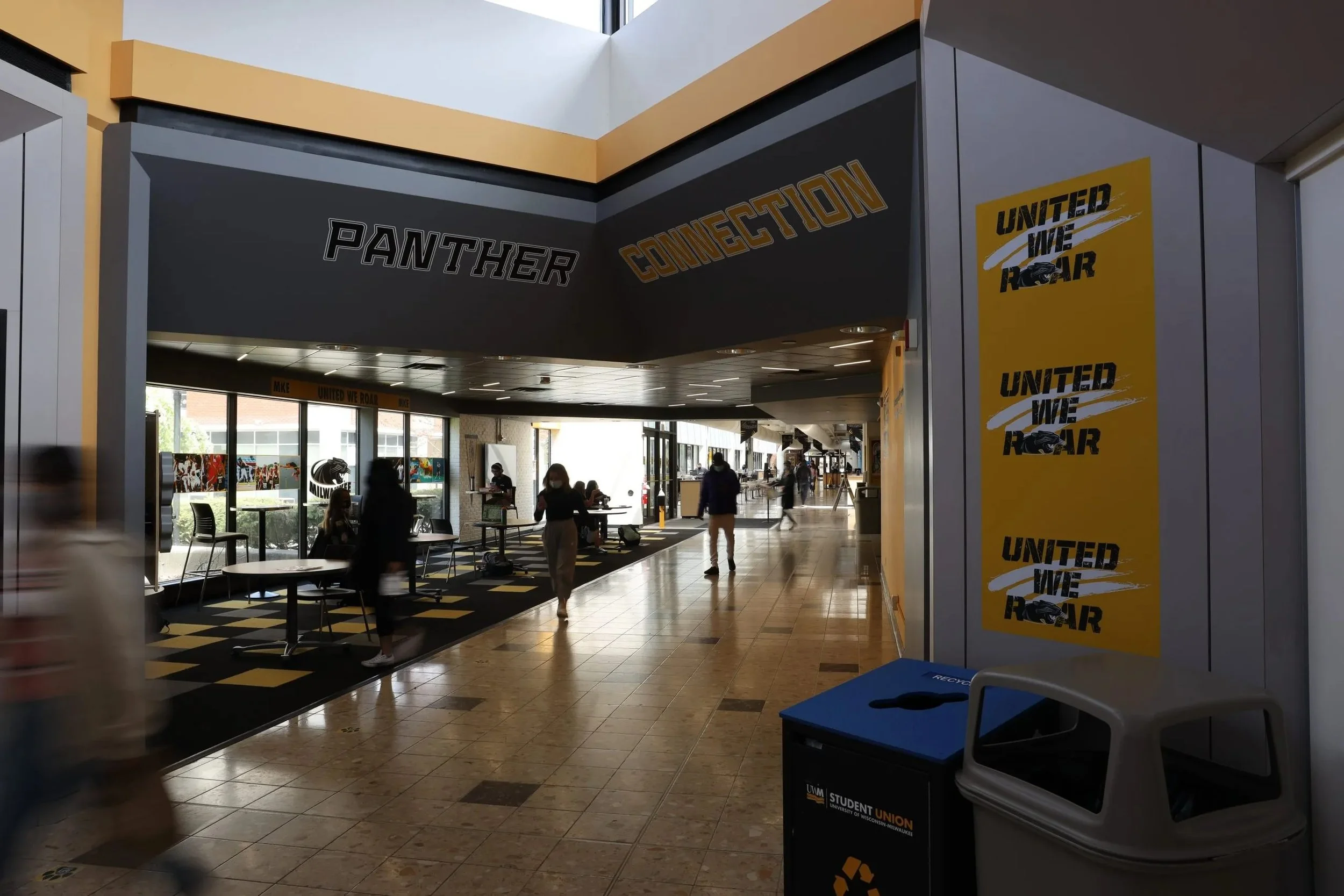 Interior of a modern public space, possibly a university or library, with people walking and sitting at tables. Signs display phrases like 'Panther Connection' and 'United We Roar'. Large windows let in natural light.