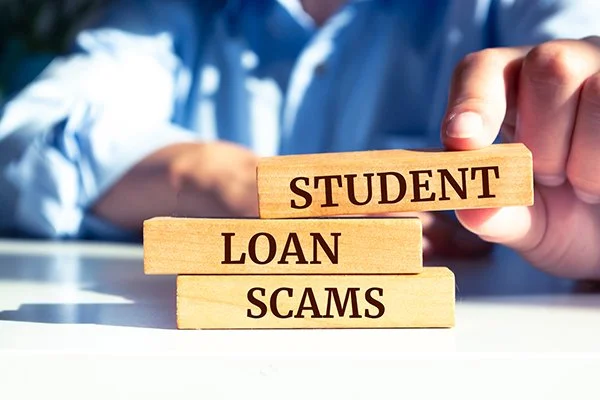 Wooden blocks with the words "STUDENT LOAN SCAMS" on a white surface, with a person in a blue shirt stacking the blocks.