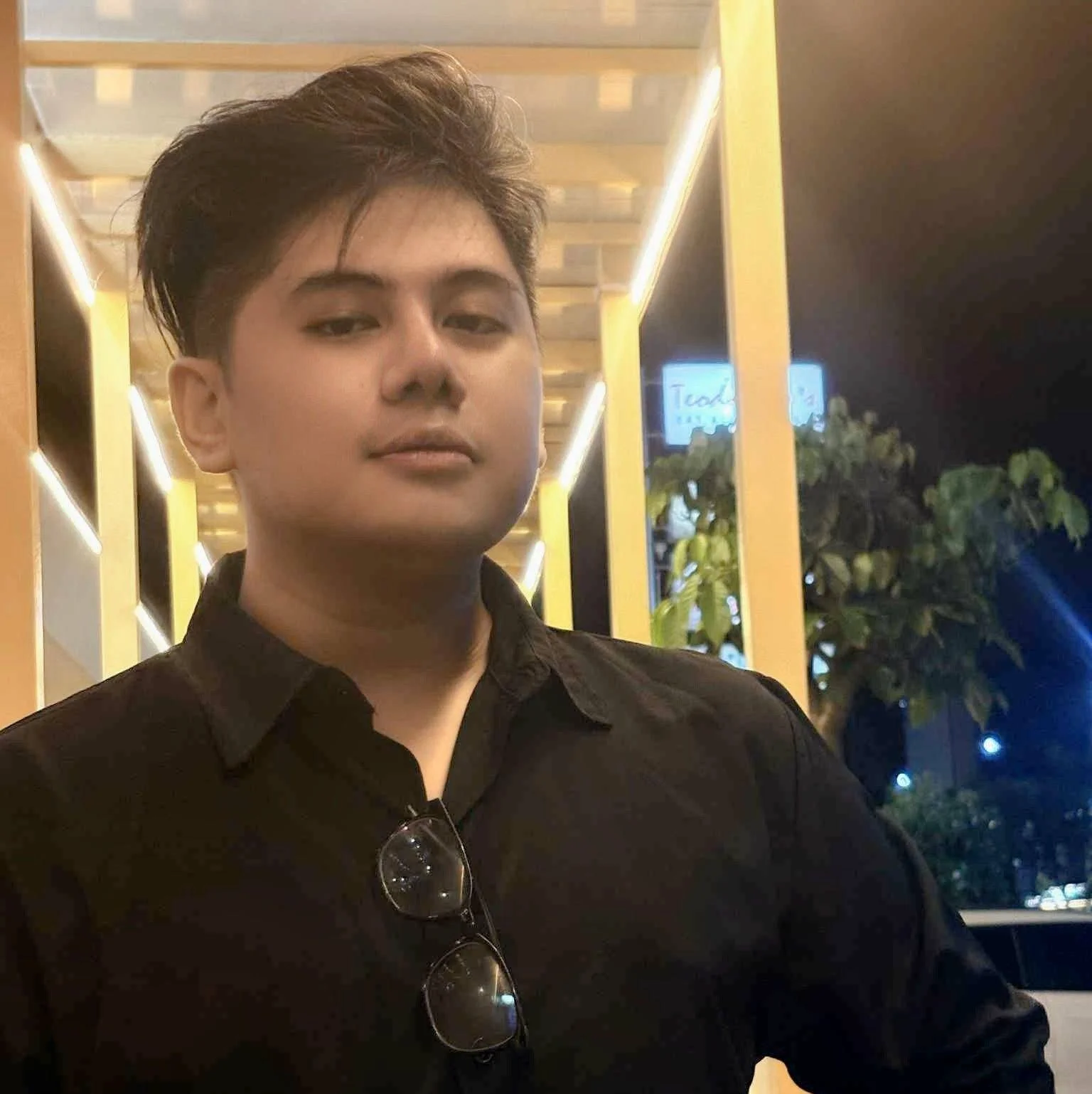 A young man with brown hair wearing a black shirt stands outdoors at night, with a yellow building with lighting, greenery, and a sign in the background.