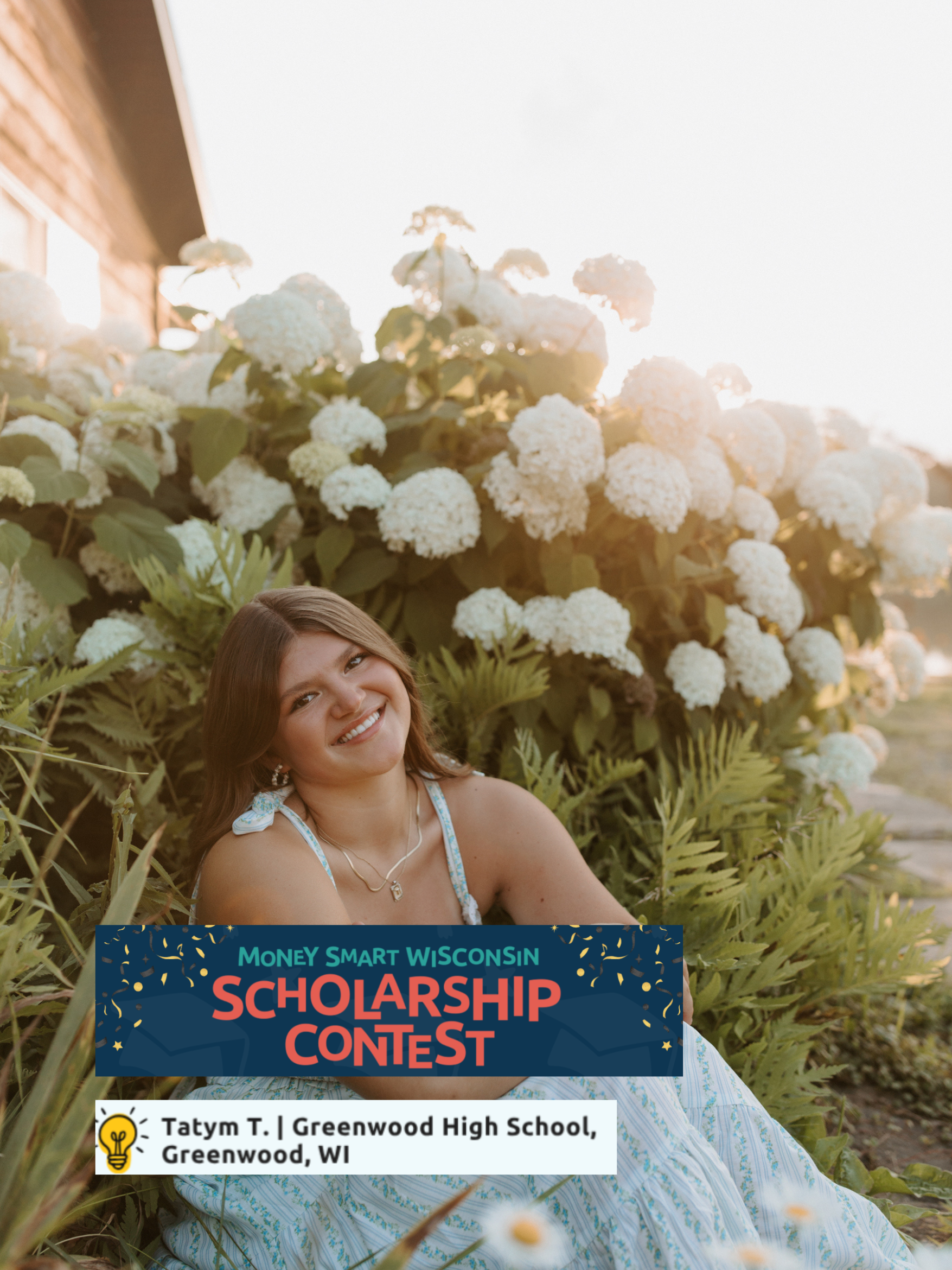 A young woman sitting in the grass outdoors with a backdrop of white hydrangea flowers, smiling at the camera. She is wearing a white and blue striped dress, holding a signed sign for a scholarship contest. The sun is shining brightly in the clear sky.