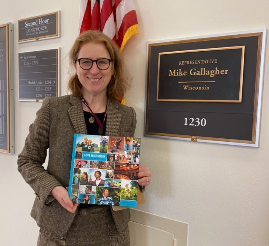 A woman holding a colorful magazine stands in front of a nameplate on a wall. The nameplate reads "Representative Mike Gallagher, Wisconsin, 1230." There are American flags behind her and other signs indicating room numbers and building information.