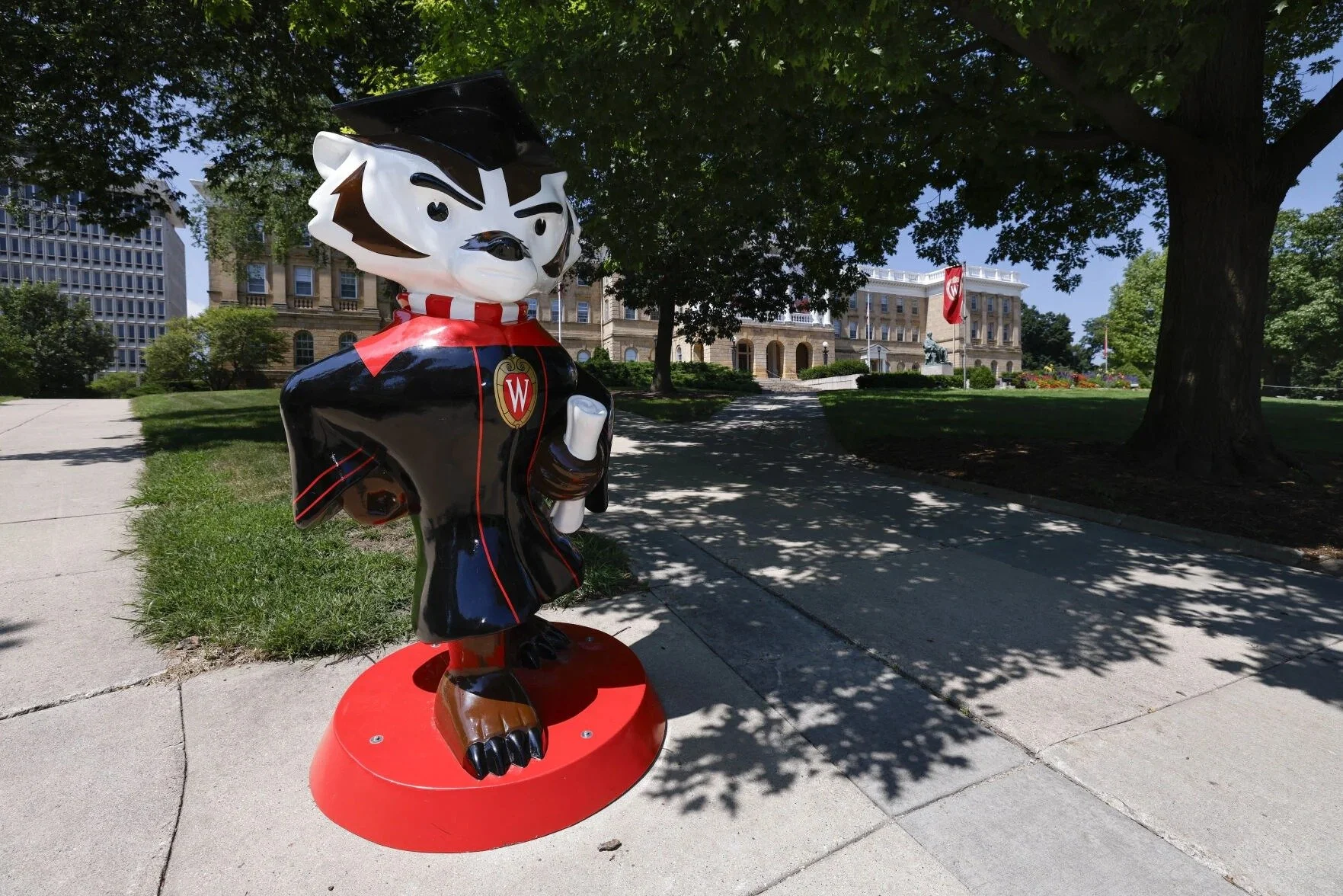 A statue of a cartoon dog dressed as a graduate, holding a rolled diploma, standing outdoors on a red circular platform, with trees and an academic building in the background at the University of Wisconsin.