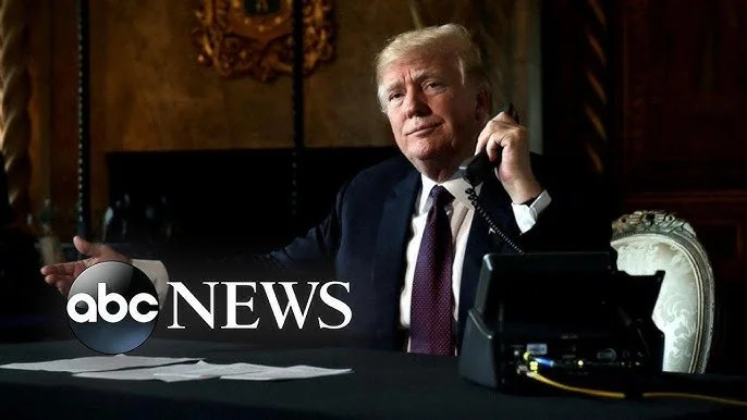 Donald Trump sitting at a desk, talking on the phone, with papers in front of him, in a dimly lit room.