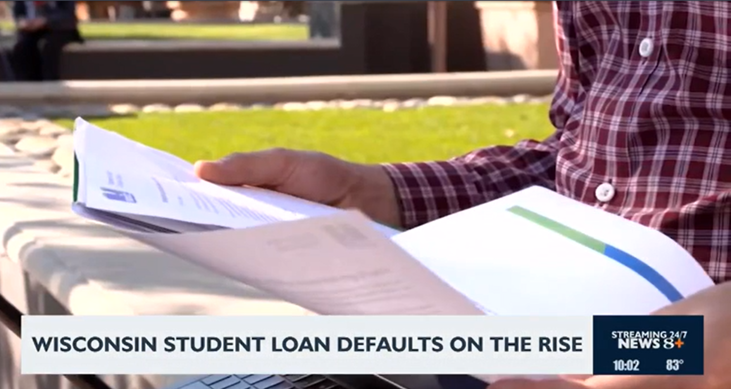 Close-up of a person in a red plaid shirt holding papers, with a news headline about Wisconsin student loan defaults on a news broadcast. DebtSmarts was interviewed for this broadcast.
