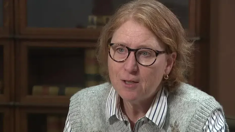 A woman with red hair, glasses, and earrings sitting indoors, wearing a light sweater over a striped shirt, in front of a wooden bookshelf.