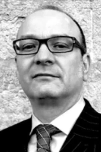 A black and white portrait of a man wearing glasses, a suit, and a tie, standing in front of a textured wall.