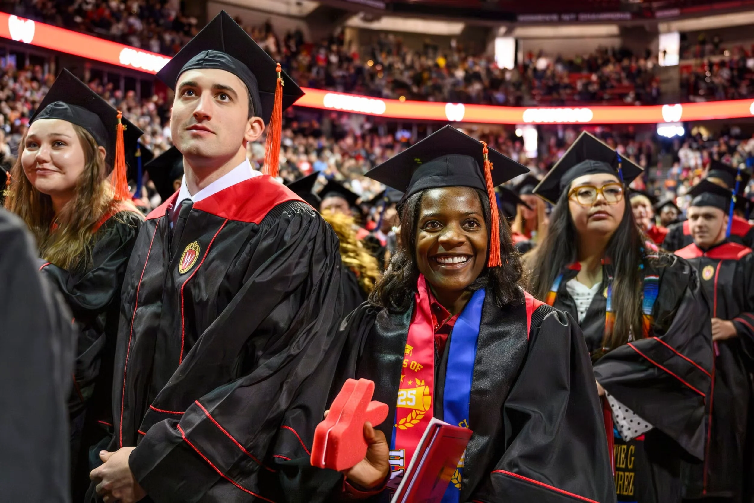 Students in academic regalia at graduation ceremony in an indoor stadium filled with attendees, holding diplomas with one smiling woman in the center wearing medals.